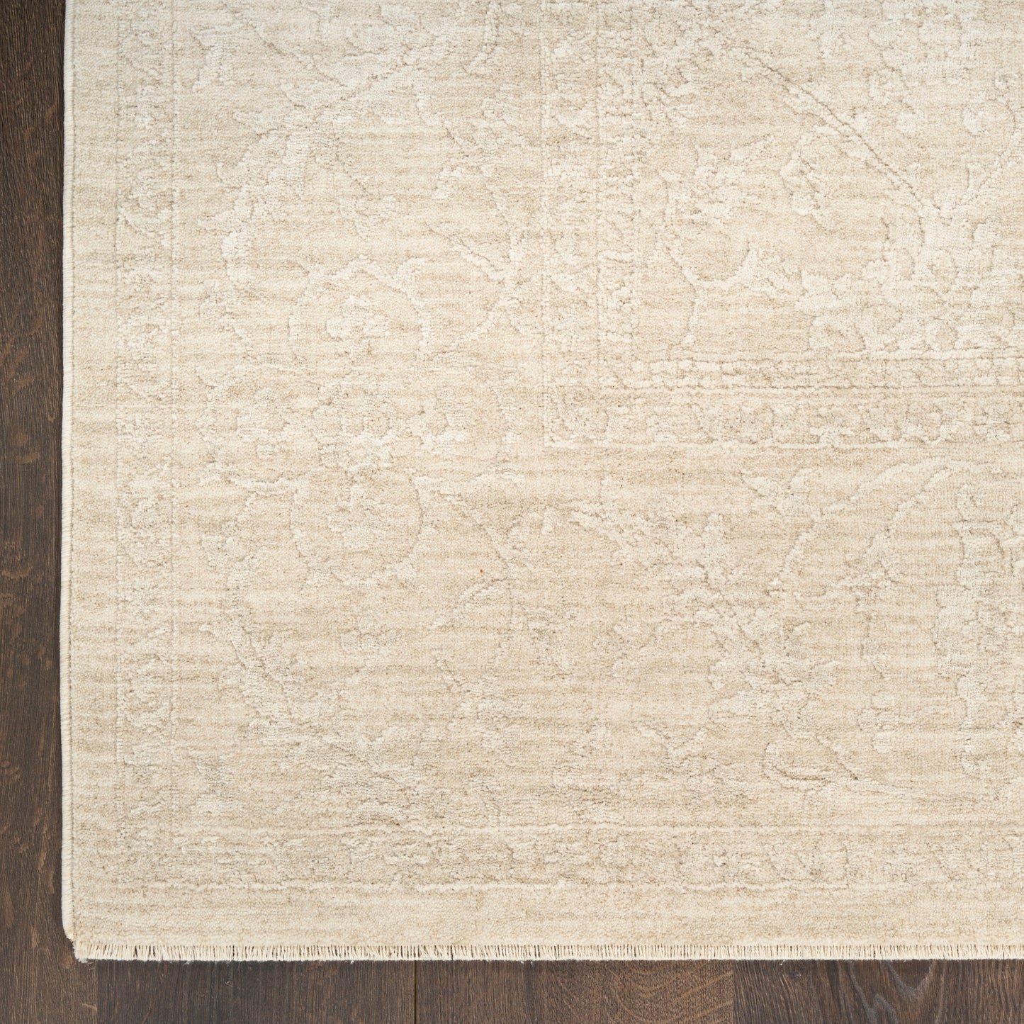 Beige patterned area rug with a subtle floral motif and fringe border on a dark wood floor.