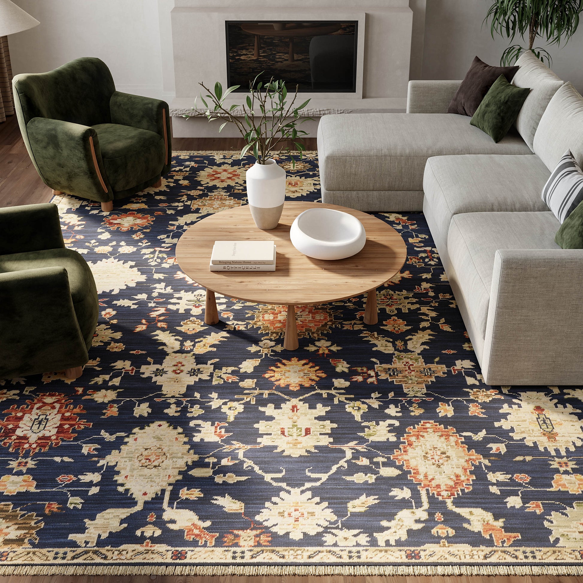 Living room with a navy floral rug, round wooden coffee table, white vase and bowl, beige sectional sofa, and green velvet armchairs.