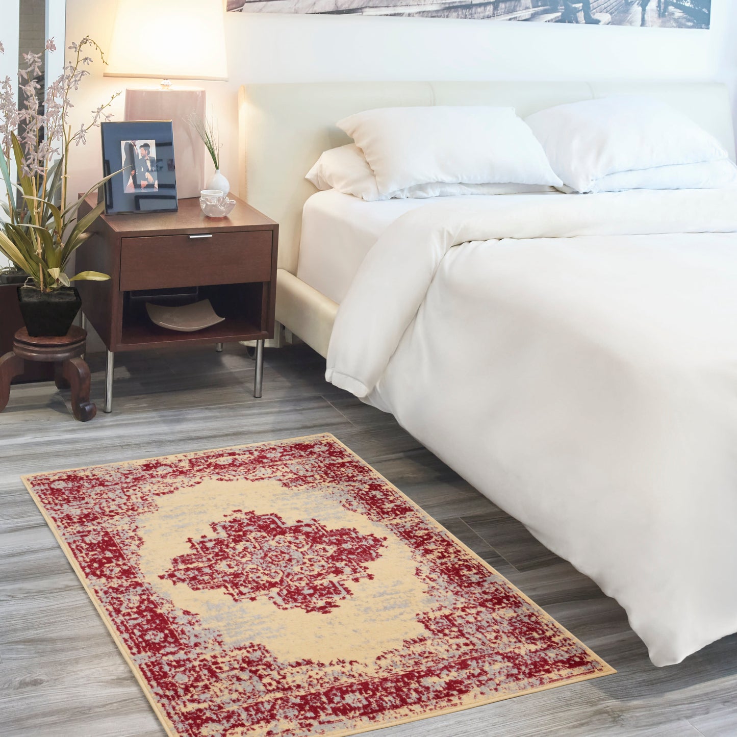 Distressed red and beige area rug with central medallion on gray wood floor beside a white bed and dark wood nightstand.