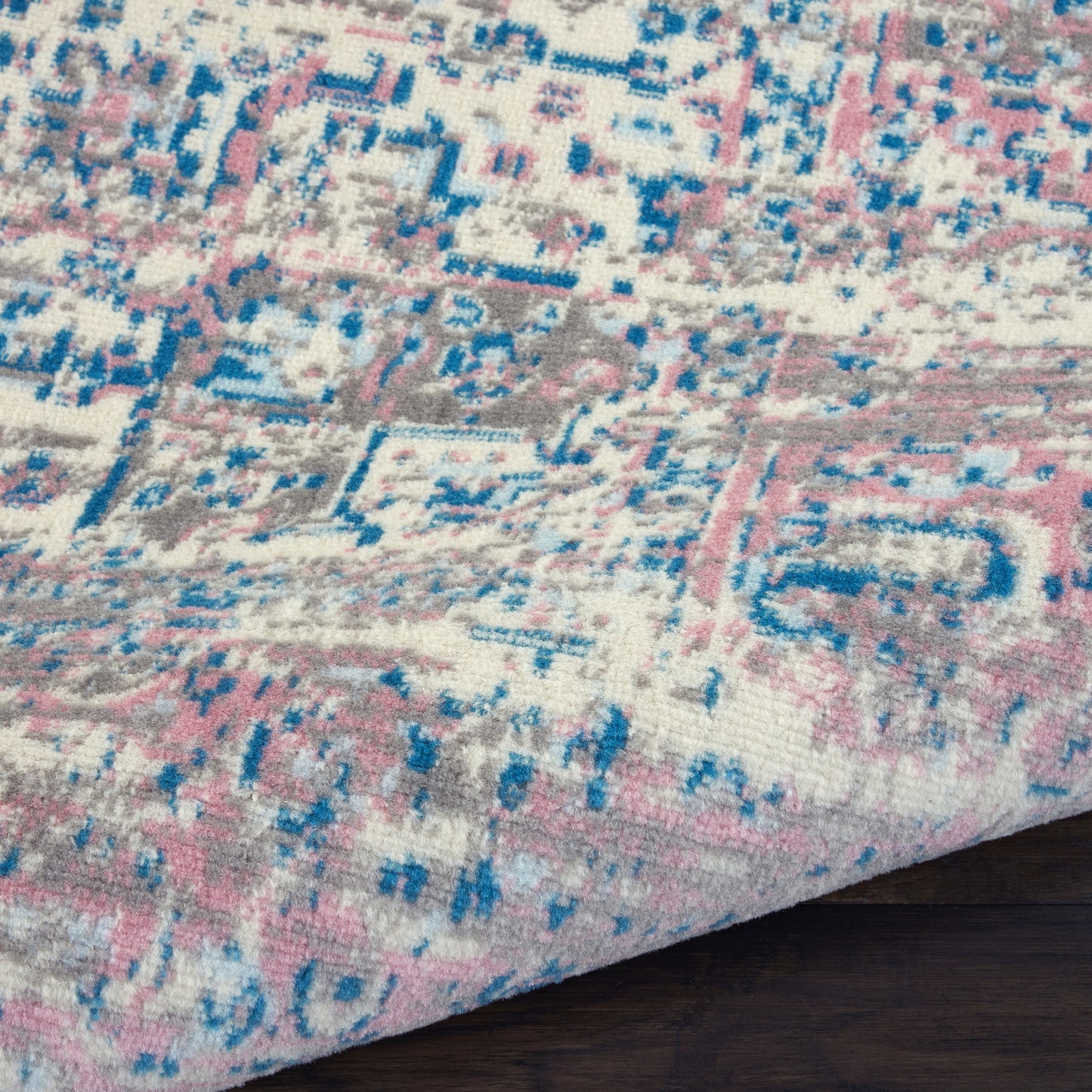 Close-up of a pastel pink, blue and gray distressed patterned rug with ivory base and a dark wood floor edge.