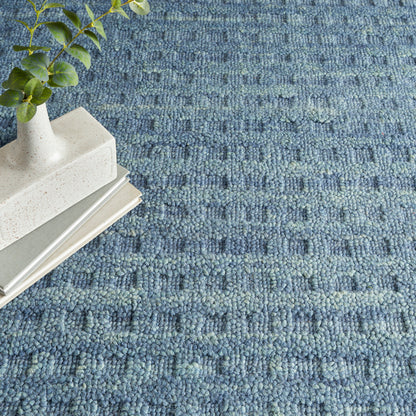 Blue woven rug with a white speckled ceramic vase and green leafy plant atop a stack of books.
