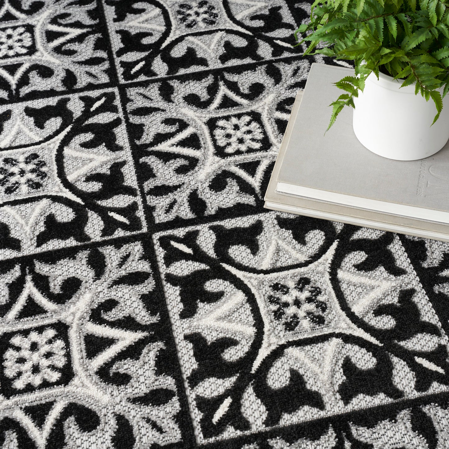 Black & white patterned rug with tile-like squares & scrolling floral motifs; plant in white pot atop stacked gray books.
