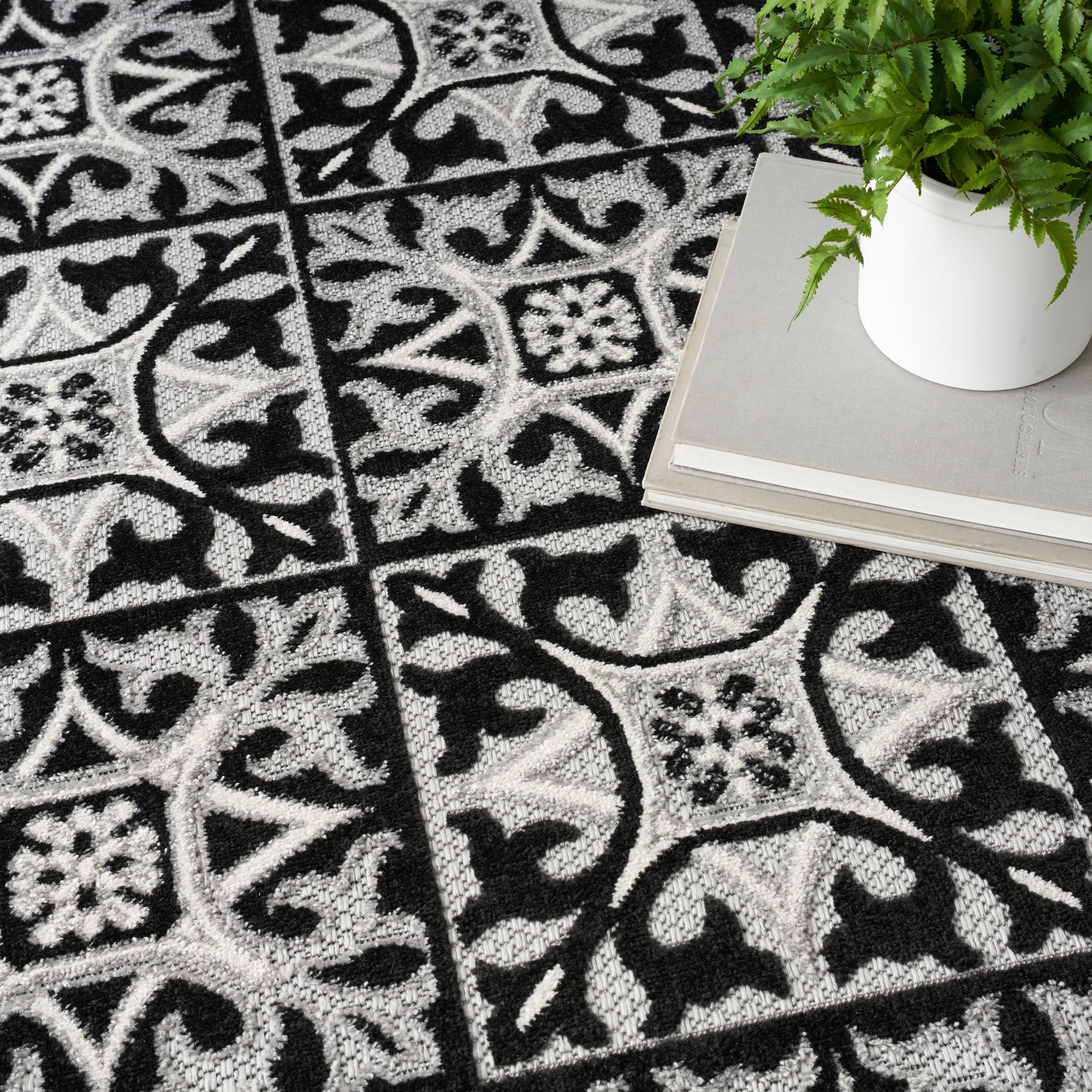 Black & white patterned rug with tile-like squares & scrolling floral motifs; plant in white pot atop stacked gray books.