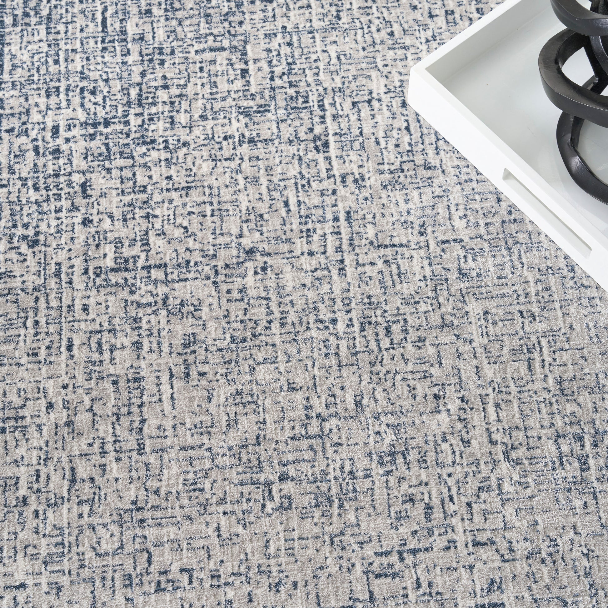 Blue and beige abstract patterned area rug with textured weave; corner shows white tray with black sculptural coil.