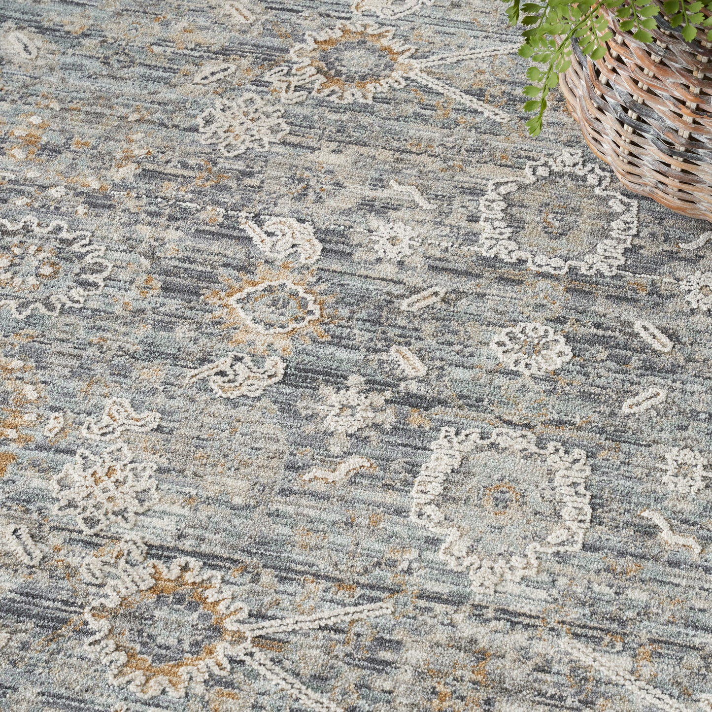 Muted blue-gray area rug with ivory floral medallions and tan accents, woven texture; wicker basket with greenery in corner.