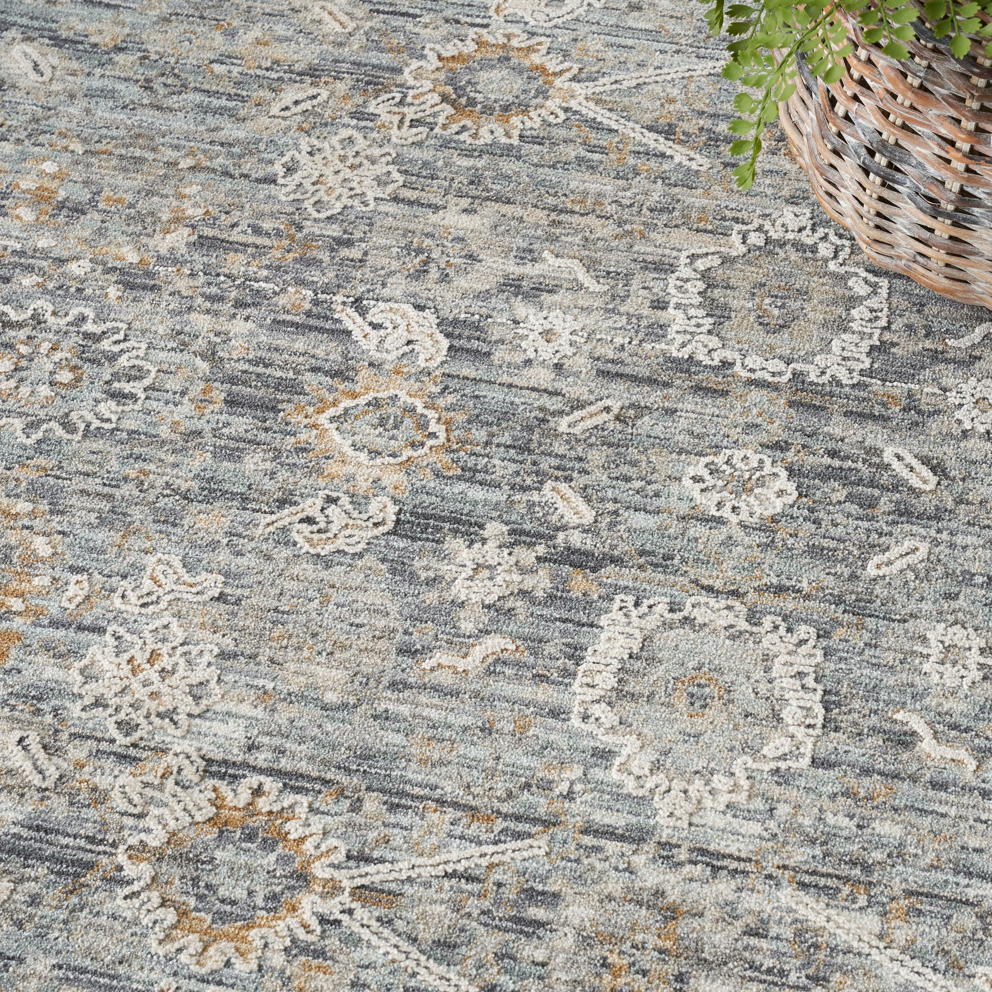 Muted blue-gray area rug with ivory floral medallions and tan accents, woven texture; wicker basket with greenery in corner.