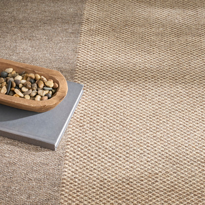 Two-tone woven rug in beige and taupe with a wooden bowl of pebbles resting on a gray book.