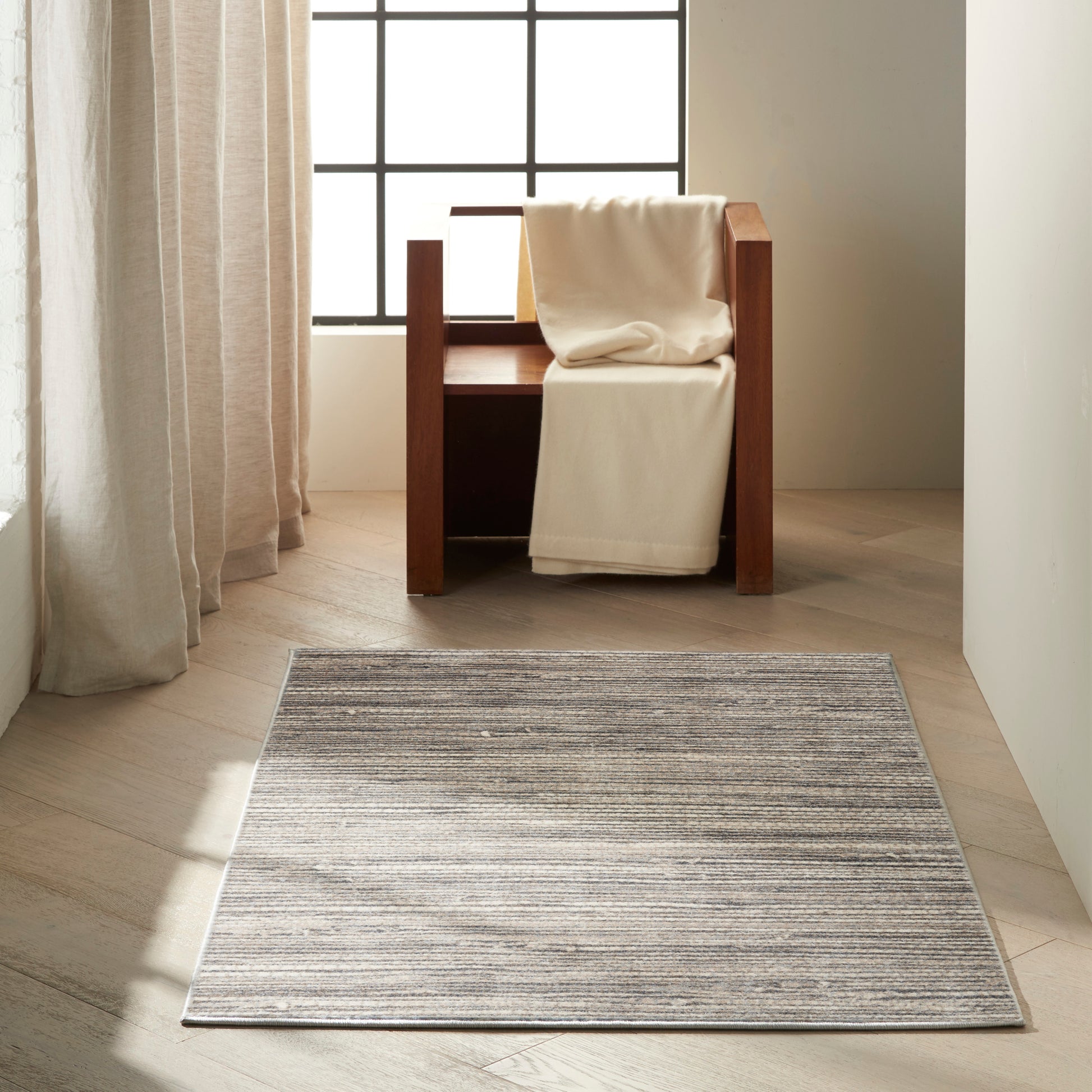 Minimalist living room with a dark wood armchair and white throw, gray striped rug, light wood floor, grid window.