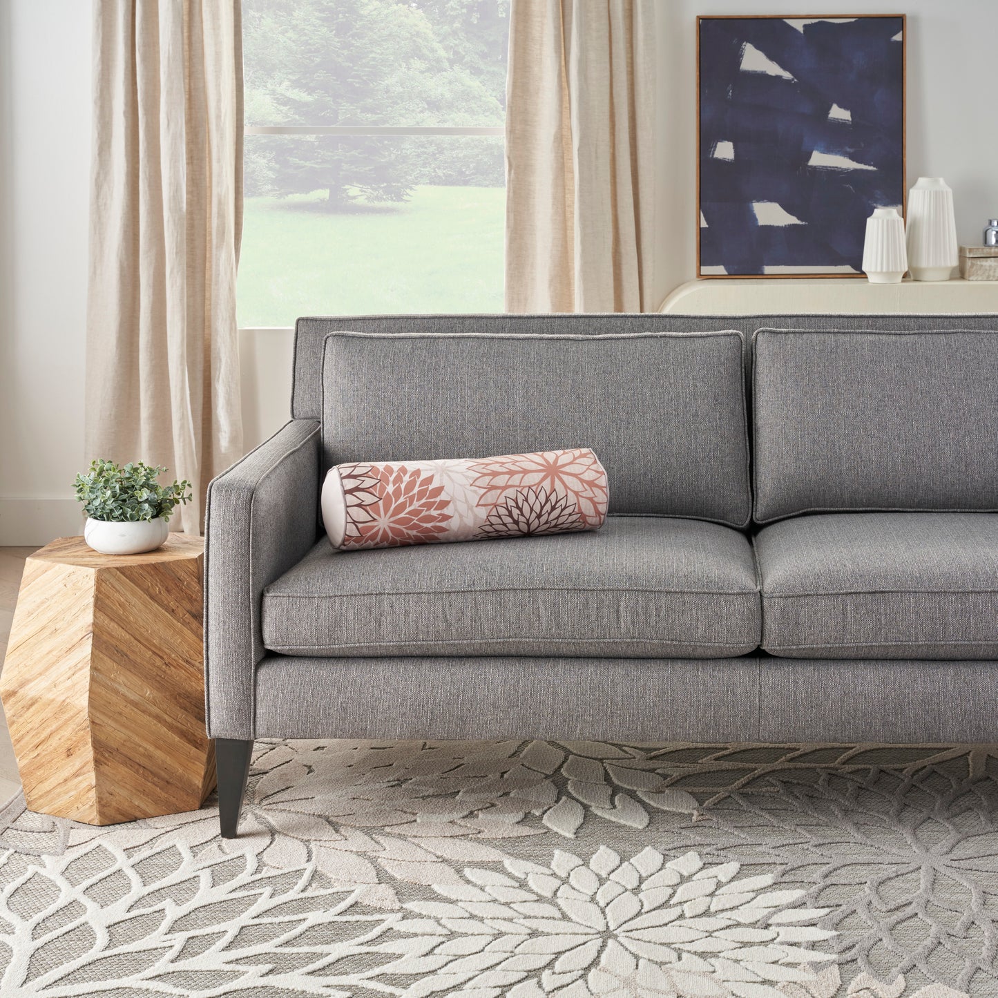 Gray fabric mid-century sofa with pink botanical bolster pillow, wooden side table, beige curtains, blue abstract wall art.