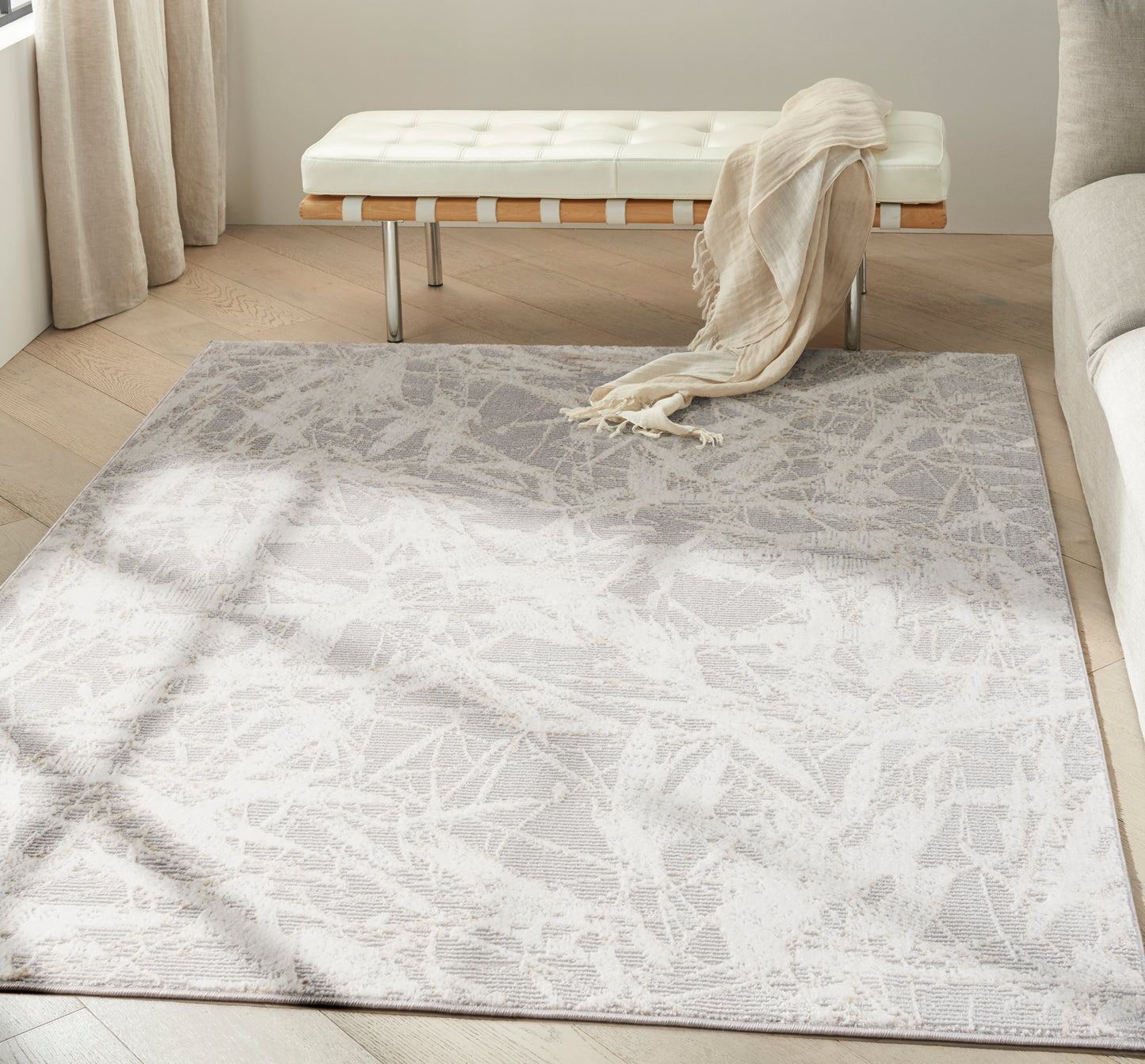 Rectangular light-gray abstract rug on wood floor; cream tufted bench with wooden slats & metal legs, beige throw draped.