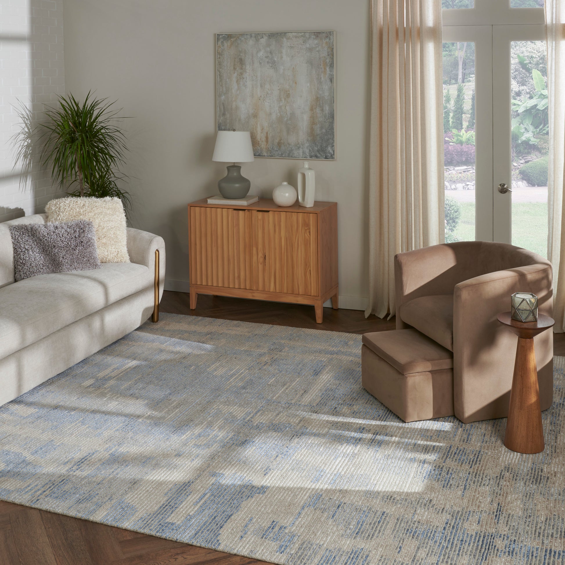 Living room with beige sofa, mid-century wooden sideboard, blue-gray rug, brown armchair and ottoman, plant, French doors.