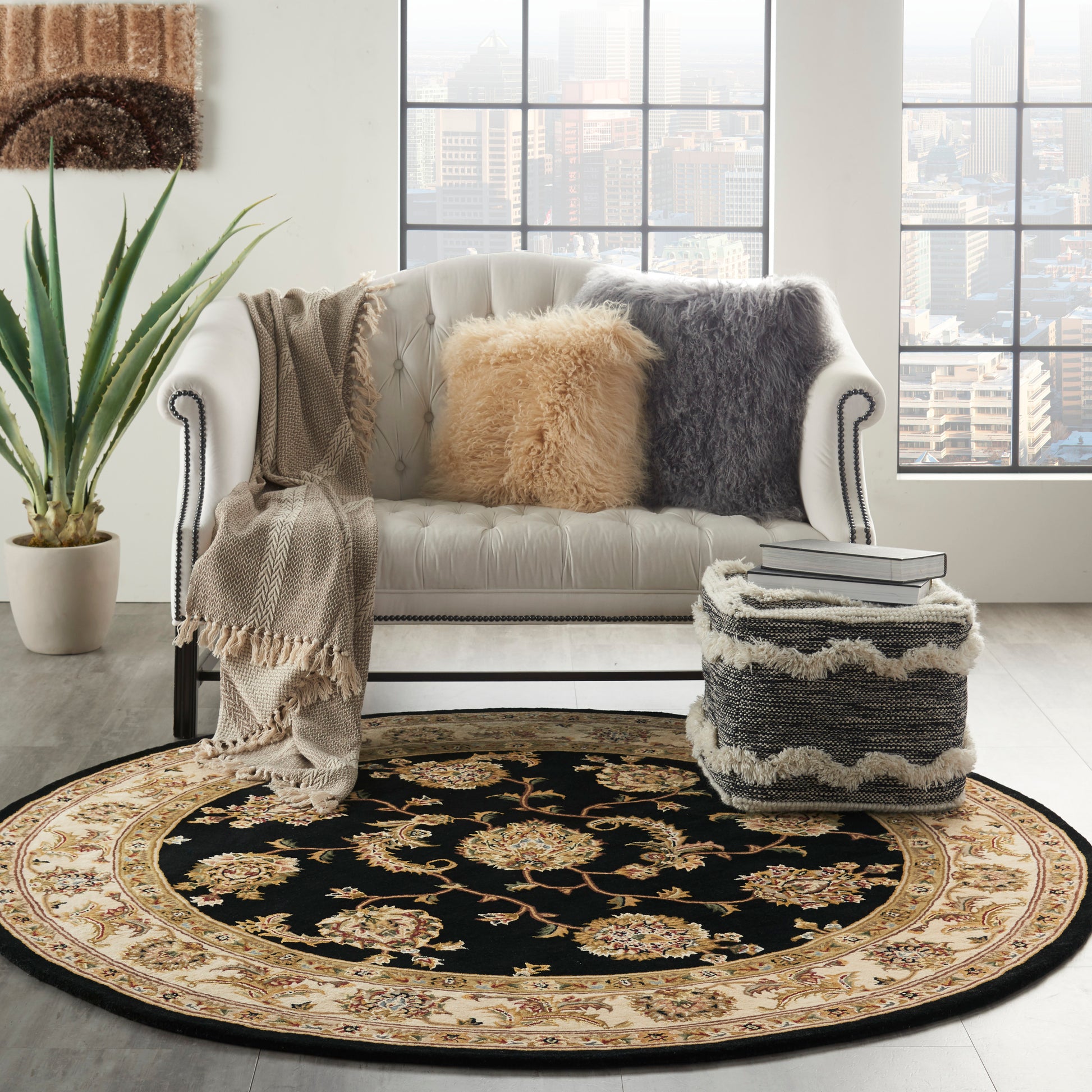 Round ornate rug, cream tufted sofa with tan and gray faux fur cushions, knitted throw, ottoman, plant, city view.