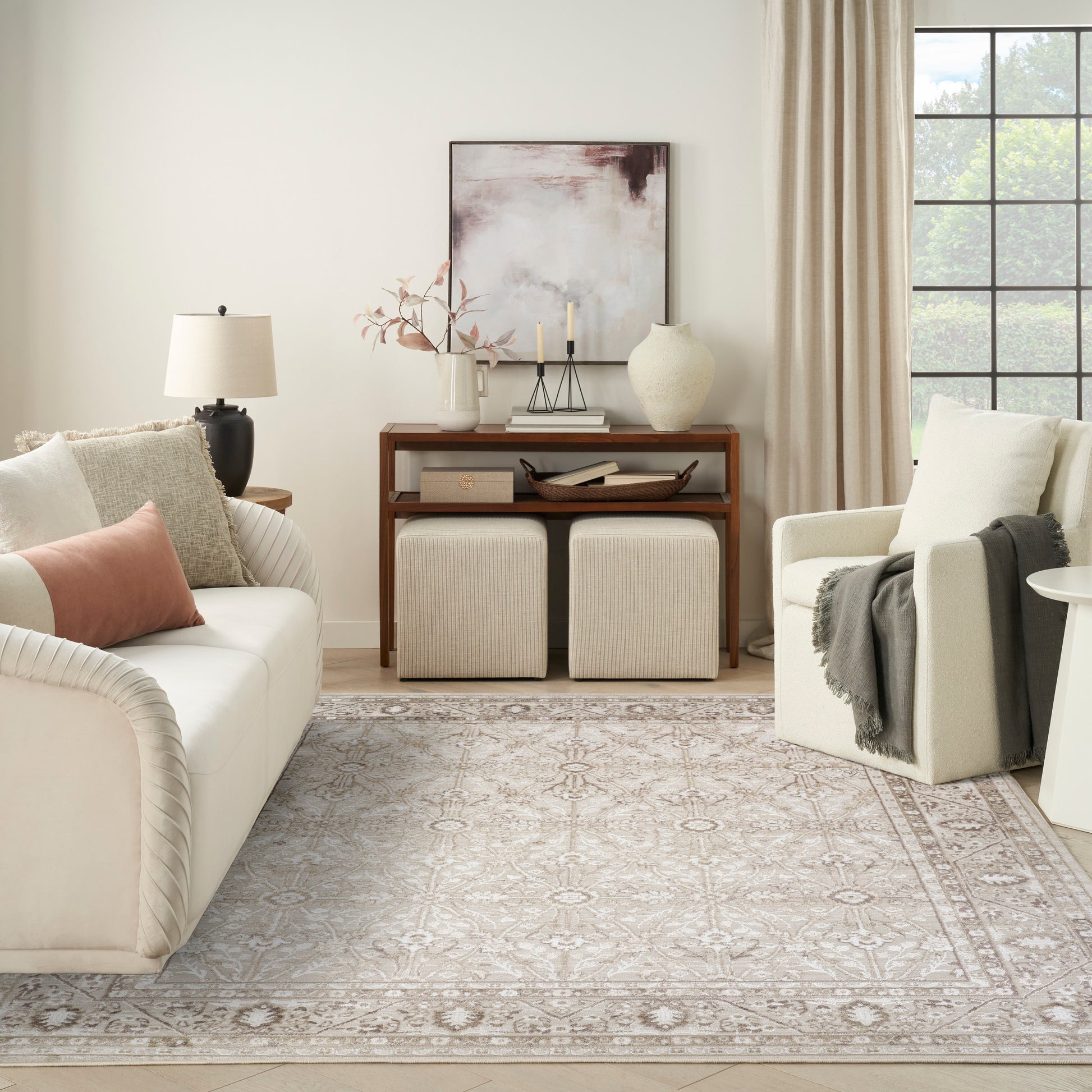 Neutral living room featuring cream sofa, pale pink pillows, beige armchair, wooden console, two ottomans, and a patterned rug.