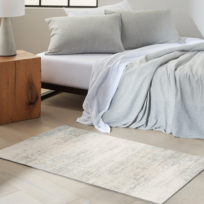Gray-toned area rug on the floor in a neutral bedroom; visible bedding includes light gray sheets, a textured gray throw and pillows, and a wooden nightstand.