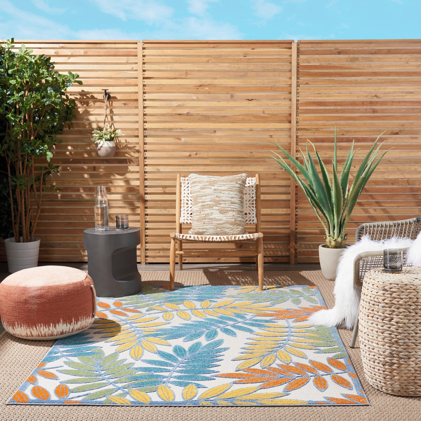 Boho outdoor patio with wooden slat fence, wicker chair and ottoman, colorful tropical leaf rug, potted plants.