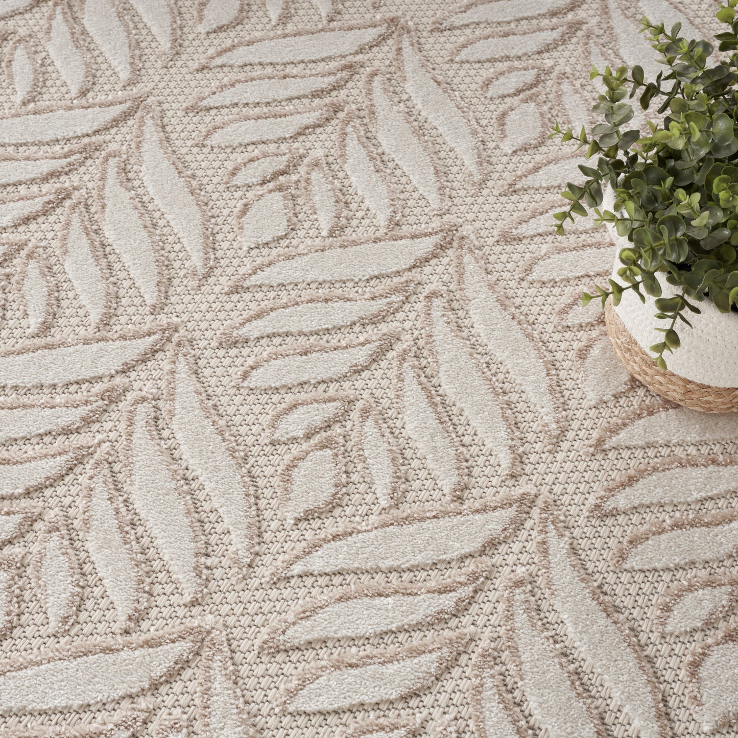 Beige leaf-patterned rug with textured pile; small potted green plant in a woven basket sits at the edge.