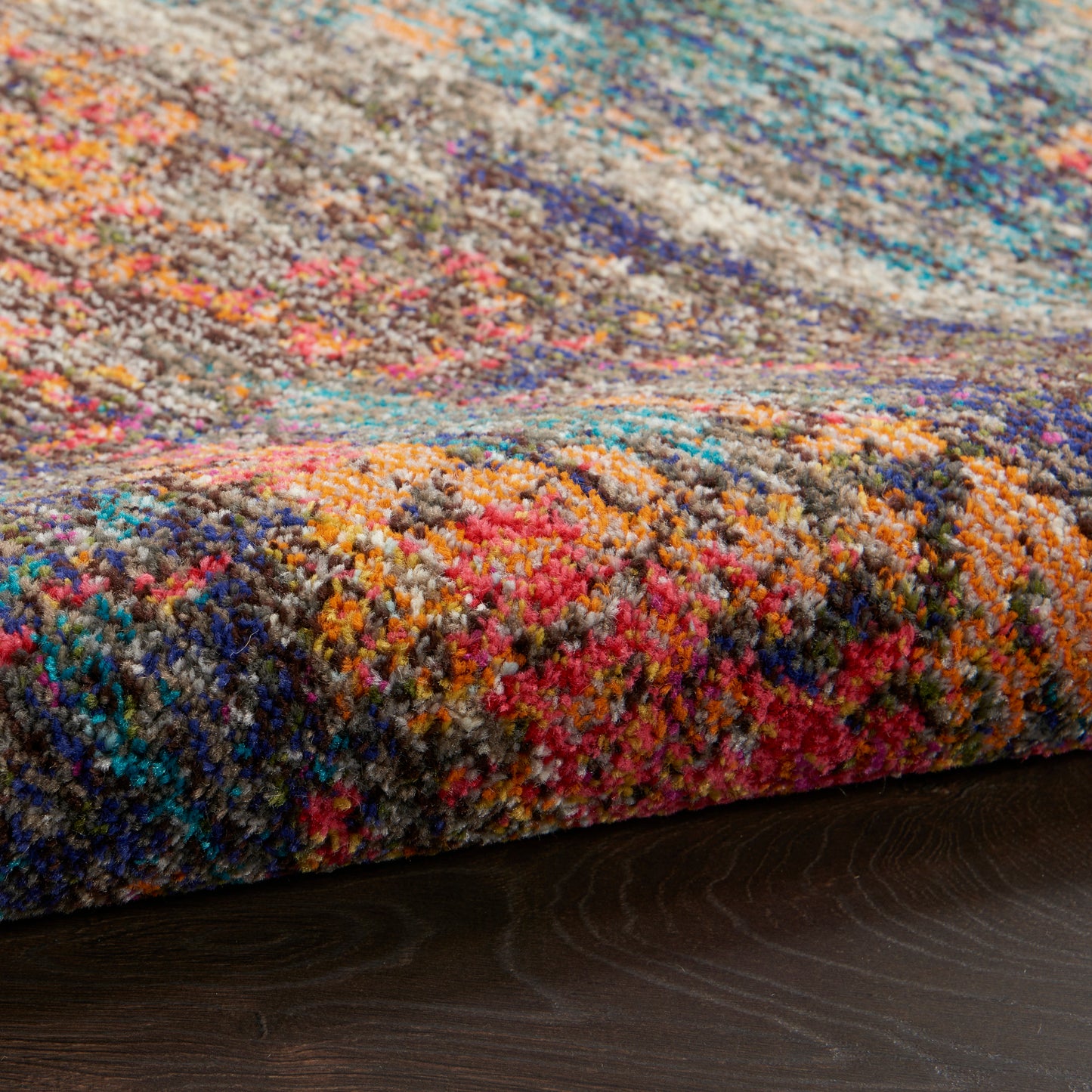 Colorful shag rug with multicolored tufted yarn in pink, blue, orange, teal and gray, edge resting on dark wood floor.