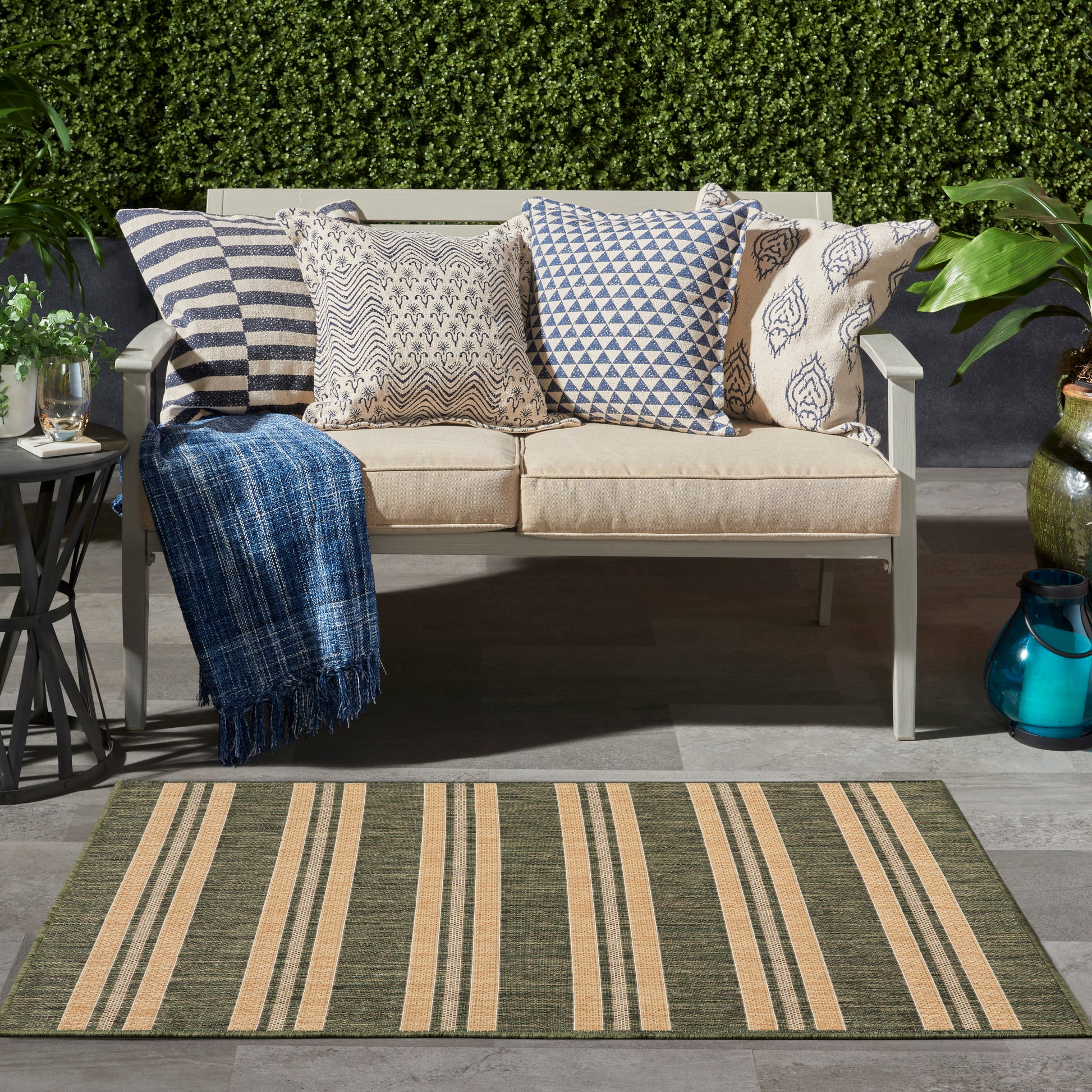 Outdoor beige sofa with blue-and-white patterned pillows and a blue throw, placed on a striped beige-green outdoor rug.