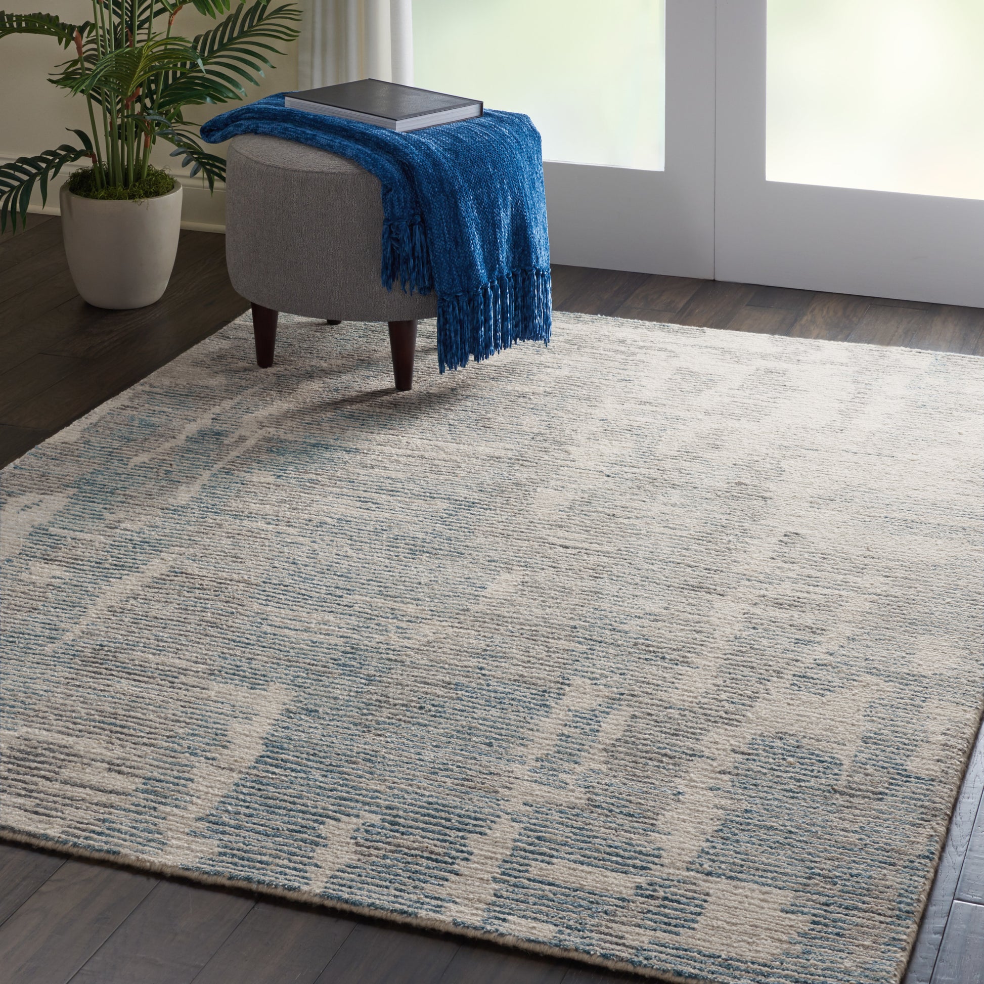 Gray round ottoman with dark wood legs, blue throw blanket and book; beige-blue patterned area rug on dark wood floor.