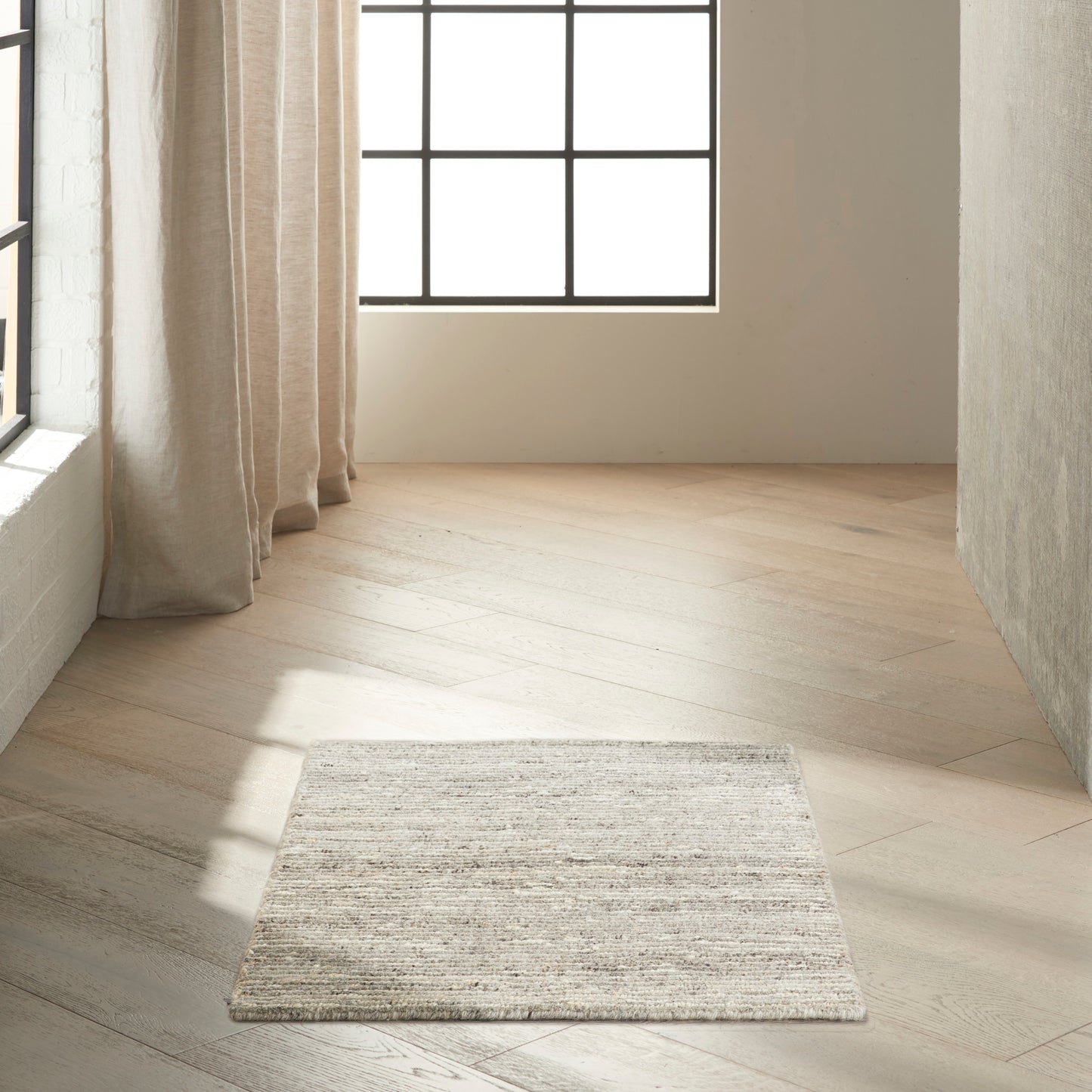 Neutral woven rectangular area rug in light gray-beige tones placed on light wood floor by a window with beige curtains.