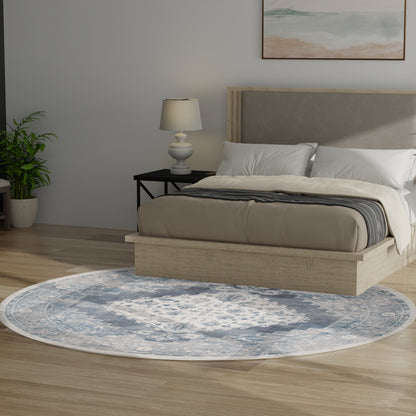 Circular blue-gray rug in a bedroom setting; beige upholstered platform bed with cream bedding & white pillows; black nightstand with lamp.
