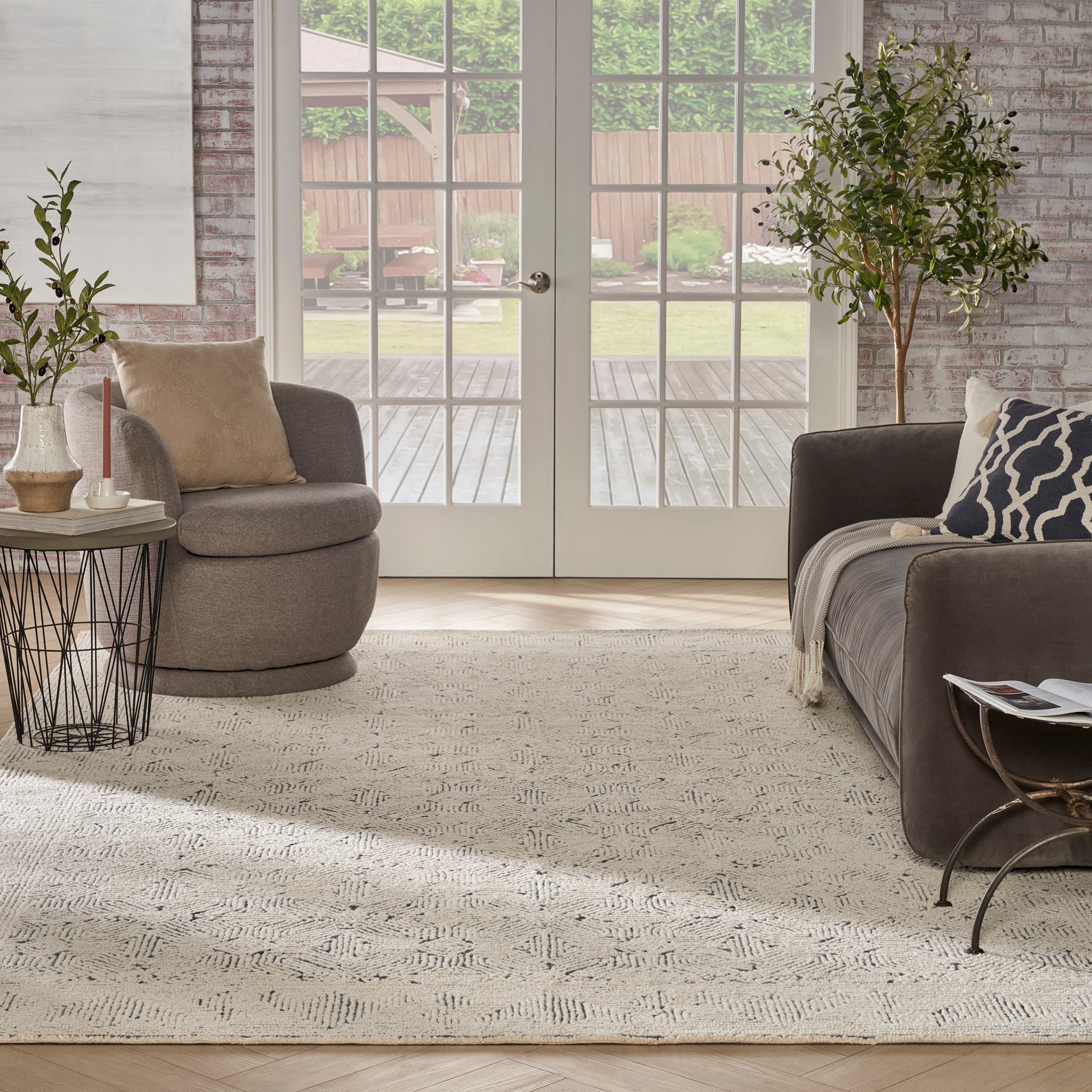 Cozy living room with gray sectional, round ottoman, beige cushions, textured rug, brick wall, potted plants, glass doors.