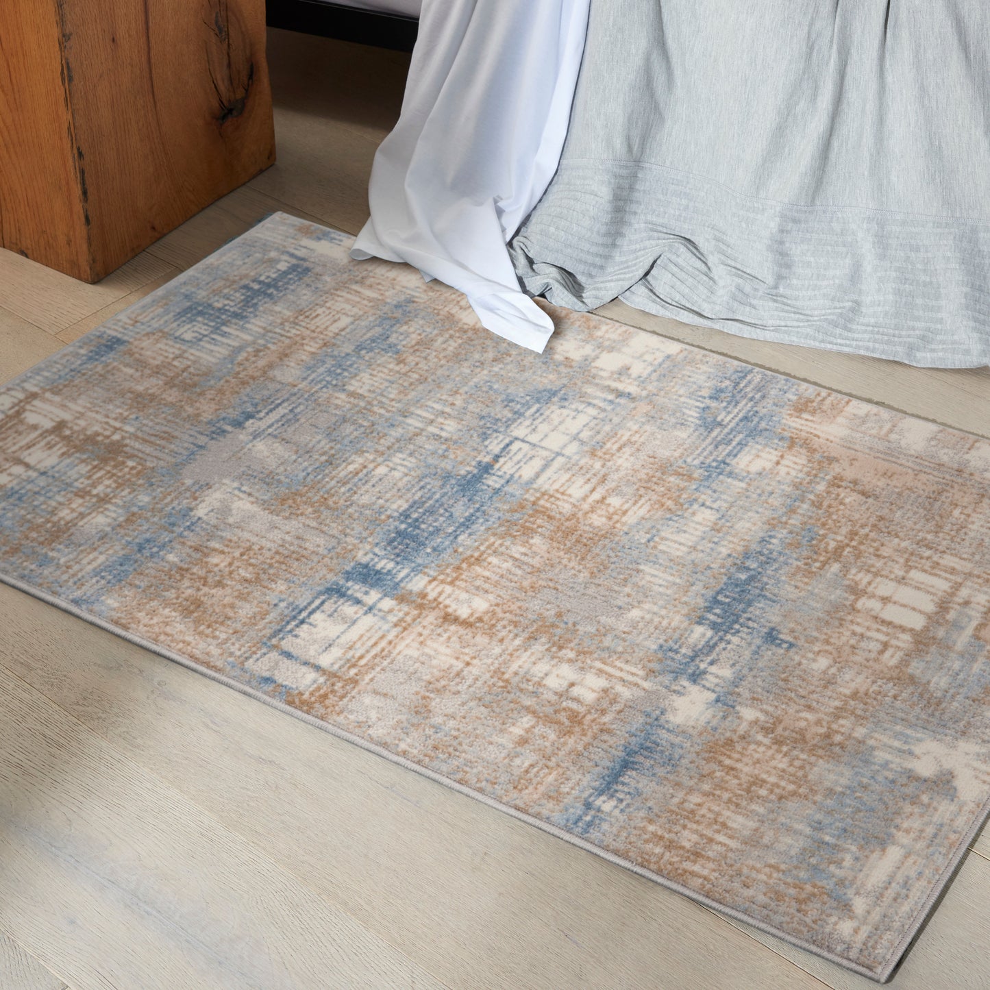 Rectangular abstract area rug in blue, tan and beige tones on light wood floor beside draped gray and white fabric.