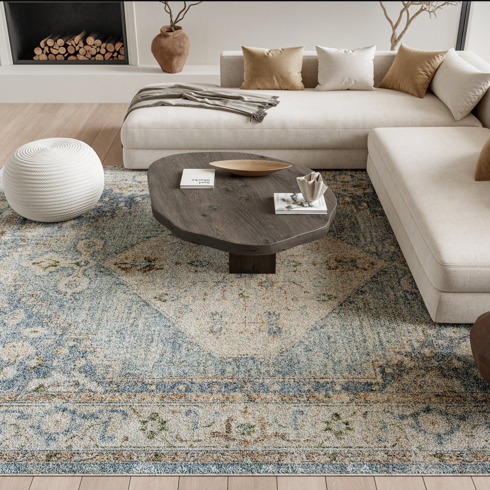 Cozy modern living room with beige sectional, dark wood coffee table, white knit pouf, blue patterned rug, tan cushions