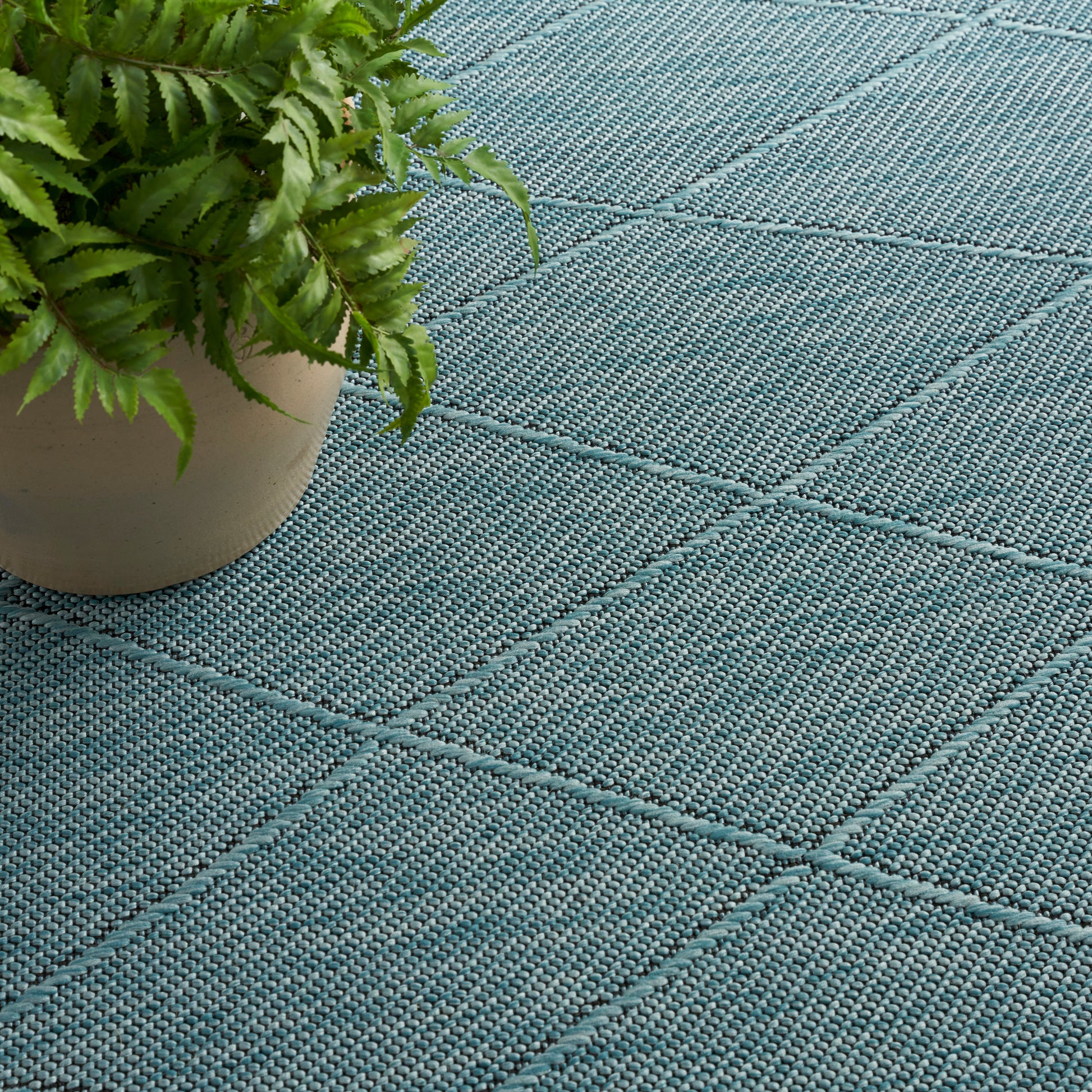 Blue-green woven outdoor rug with diamond quilted pattern; potted fern on the left.