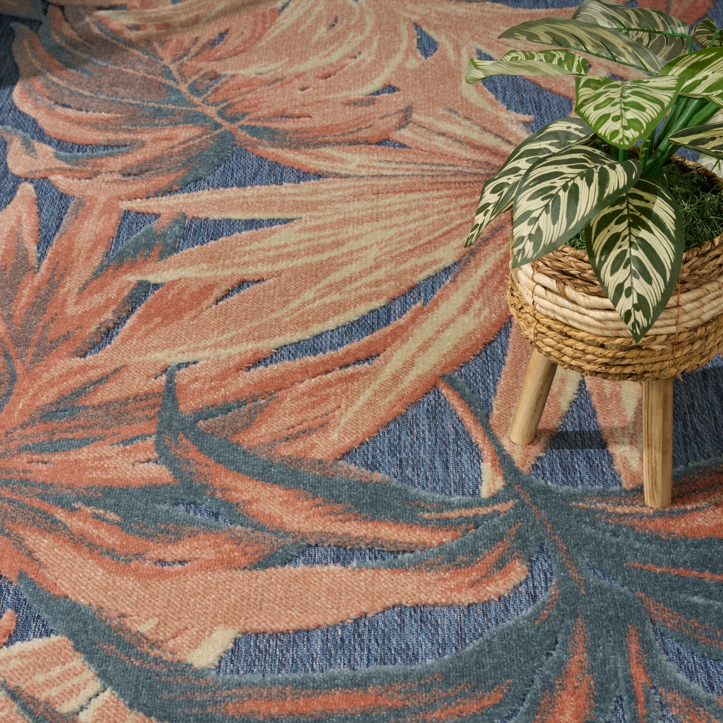 Peach and blue abstract palm-leaf rug, with a potted variegated plant in a woven basket on a wooden stool.