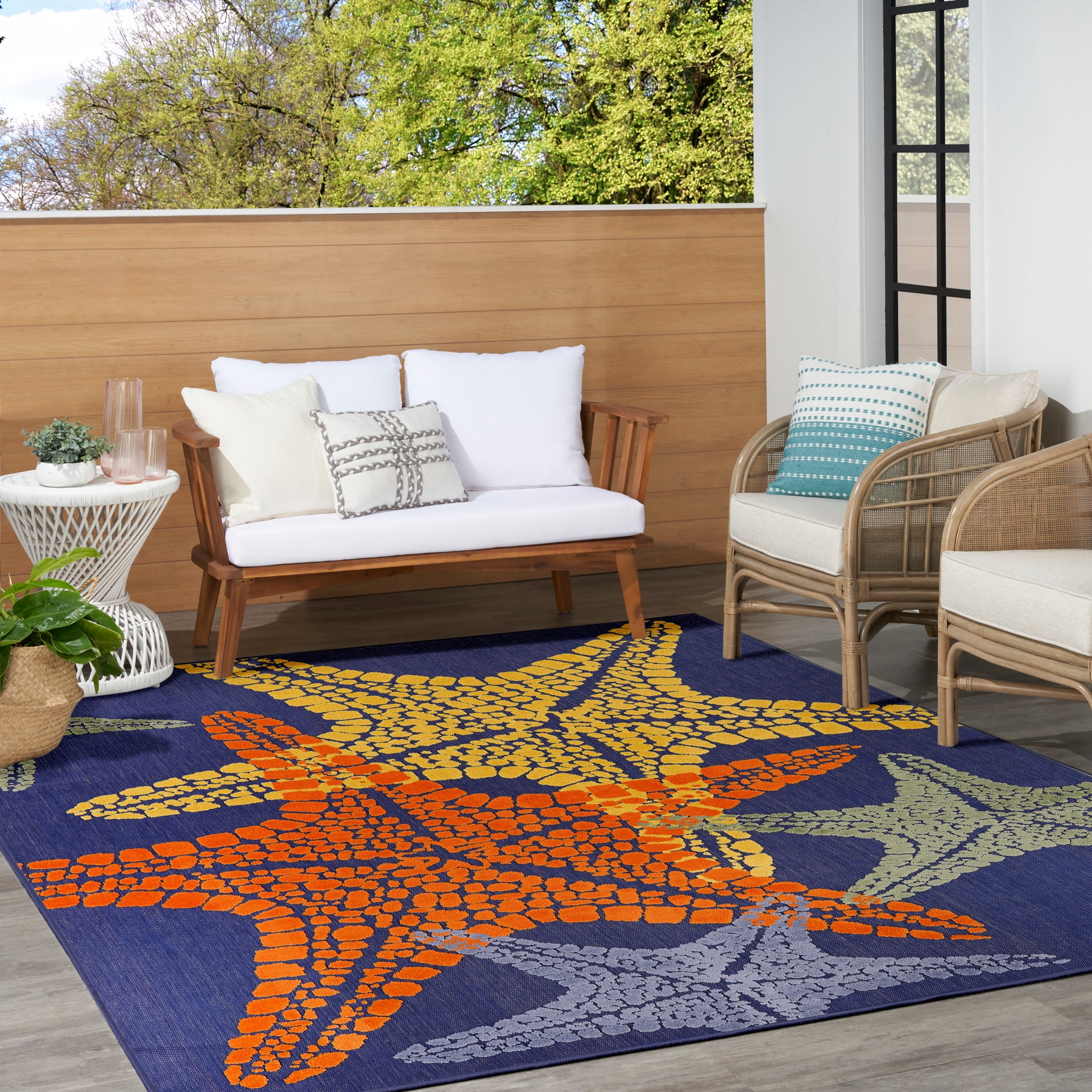 Outdoor rug with a blue starfish-pattern and orange accents on a patio, with a white-cushioned wooden sofa and wicker chairs nearby.