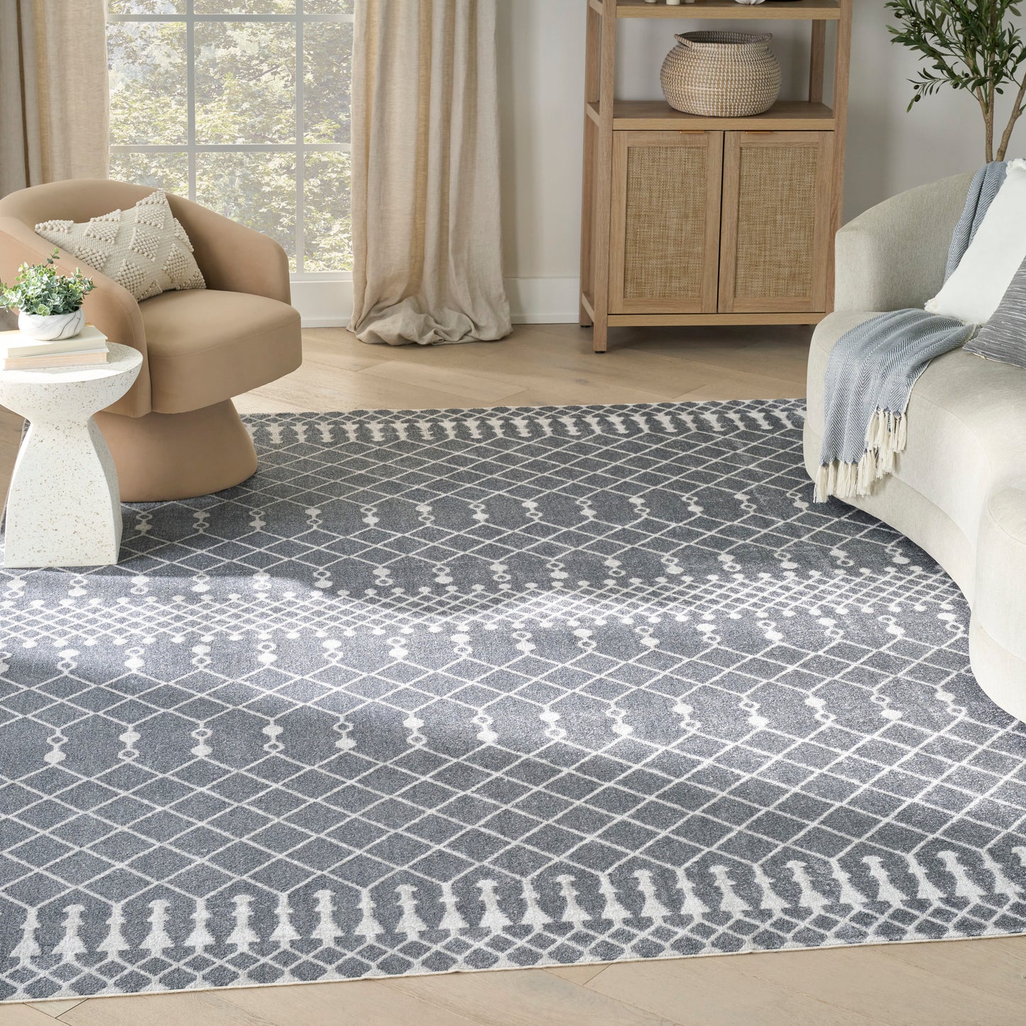 Grey and white geometric area rug in a living room with a beige curved chair, white side table, sofa, and wicker cabinet.