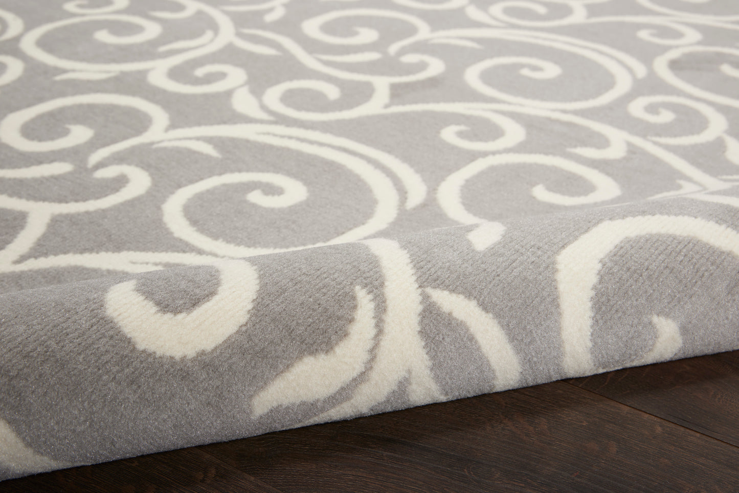 Soft gray rug with white ornate scroll pattern, plush pile on dark hardwood floor.