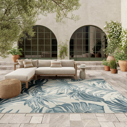 Neutral outdoor sectional sofa with wood frame, blue leaf rug, arched green-framed windows, potted plants on stone patio.