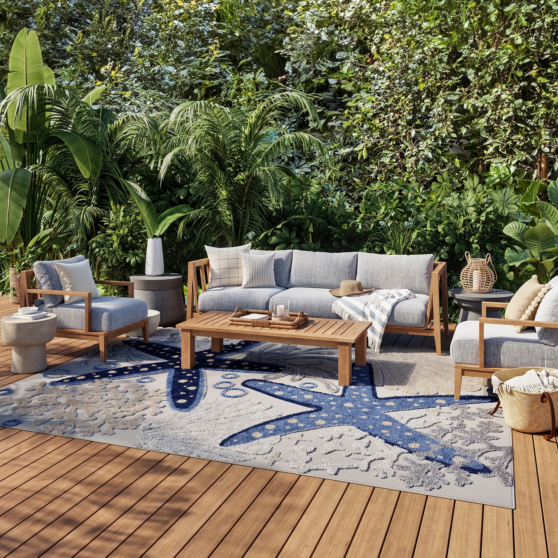 Wood-framed outdoor lounge set with gray cushions on a blue-patterned rug, atop a wooden deck amid tropical plants.