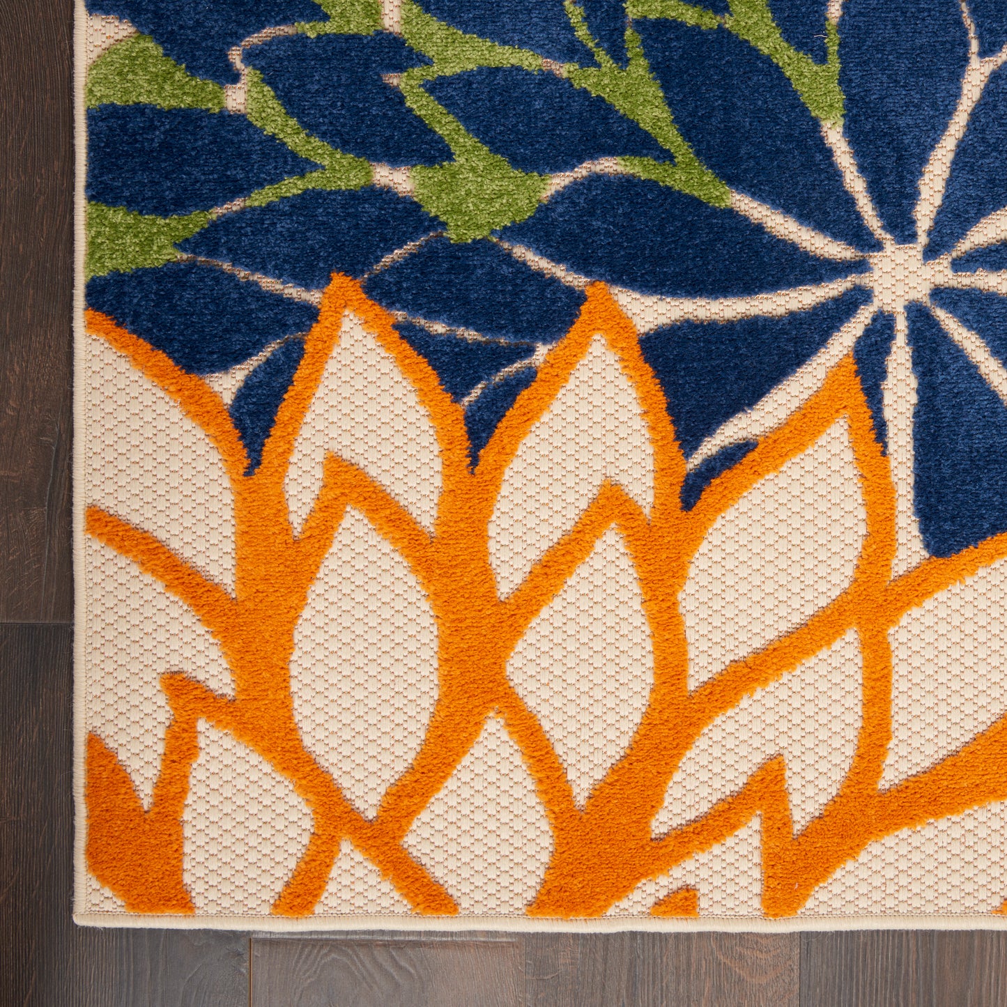 Corner of a textured flat-weave rug with bold orange leaf motifs on a white field, plus blue floral and green accents.