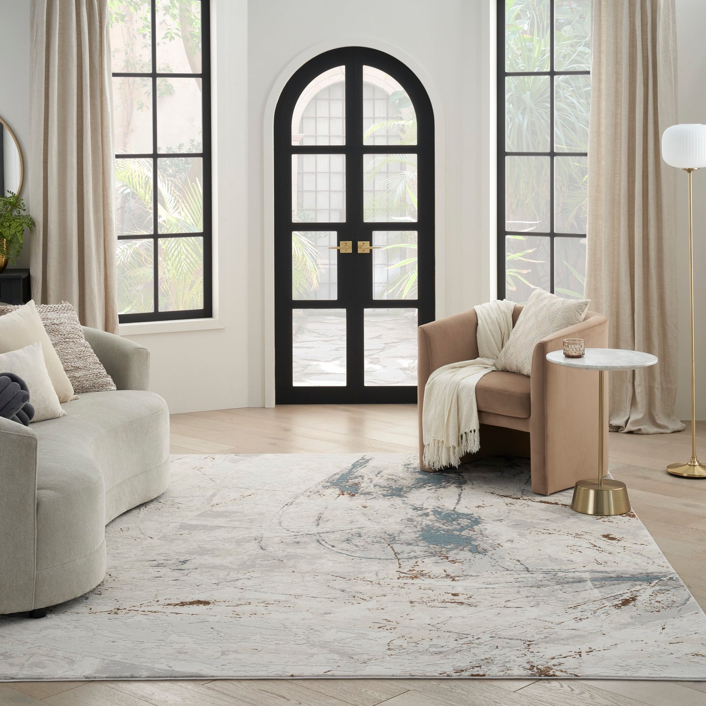 Modern living room with arched black-framed glass door, neutral sofa, tan armchair, marble side table, & gold floor lamp, blue-grey rug.