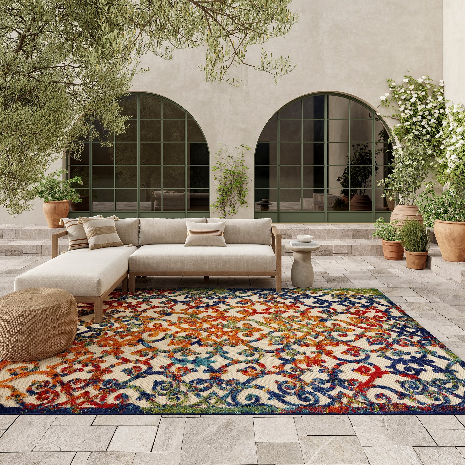 Colorful ornate rug on an outdoor patio with a cream sectional sofa, woven pouf, stone pavers, arched doors, and potted plants.