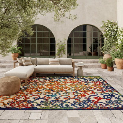 Colorful ornate rug on an outdoor patio with a cream sectional sofa, woven pouf, stone pavers, arched doors, and potted plants.