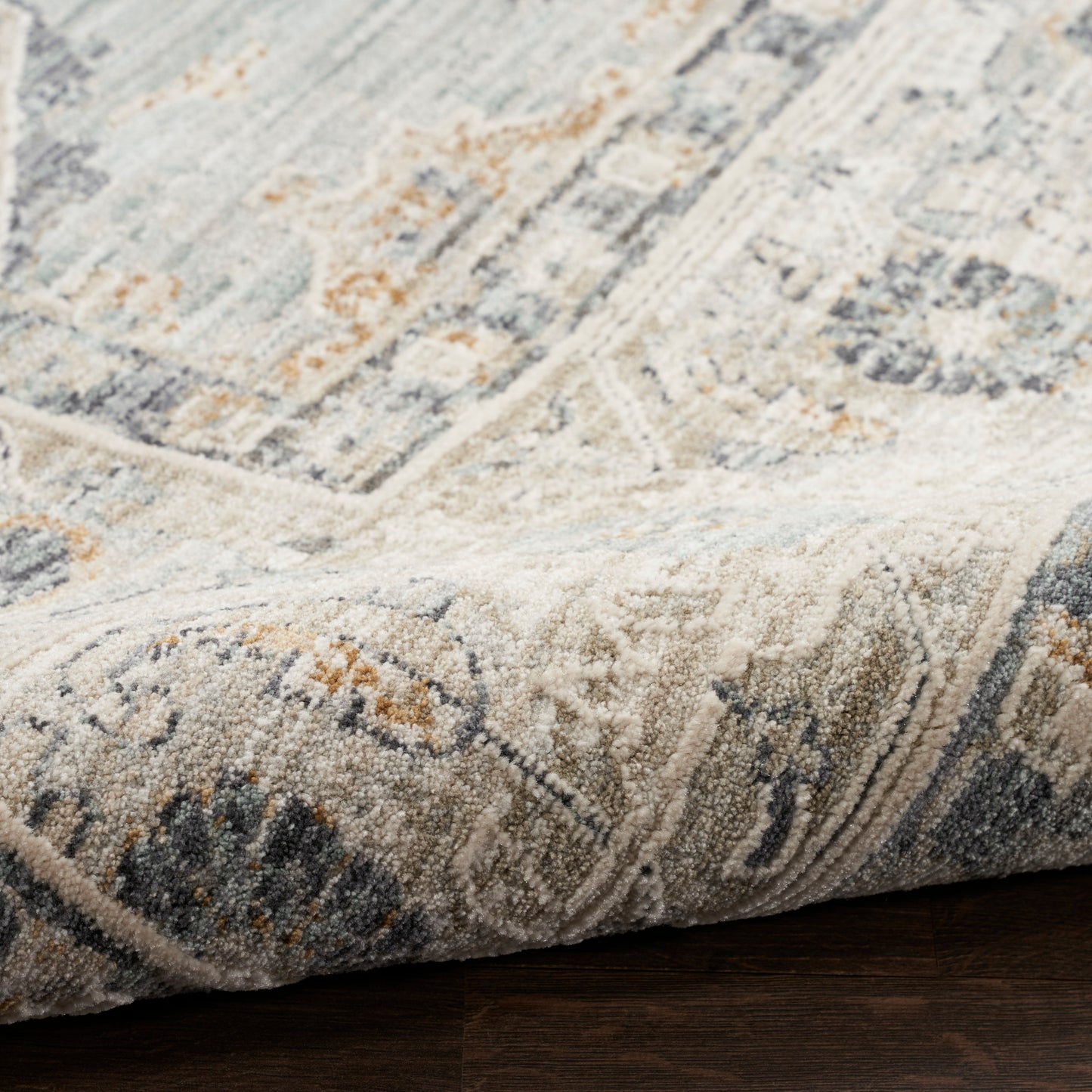 Close-up of a traditional beige-blue-gray oriental rug with orange accents, dense pile, folded corner on dark wood floor.