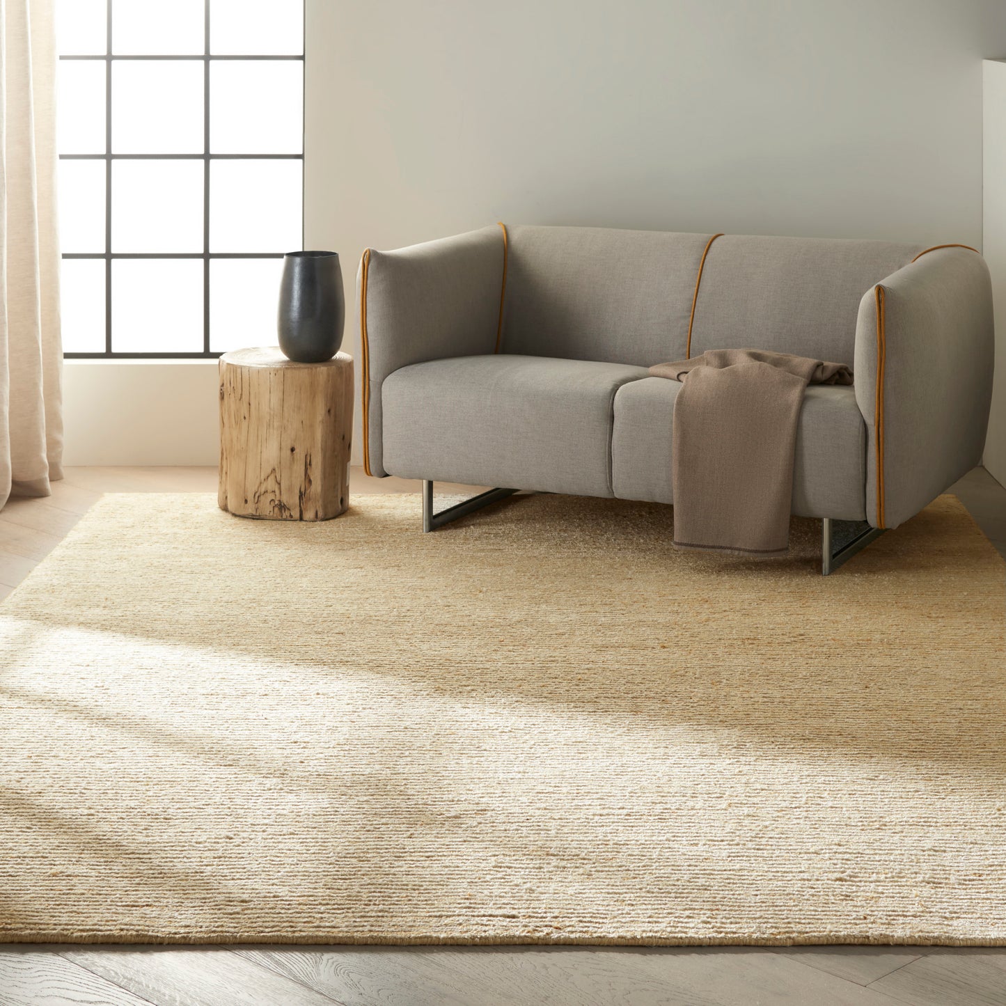 Light gray sofa with orange piping and metal legs, beige throw, on a jute rug beside a stump table with black vase