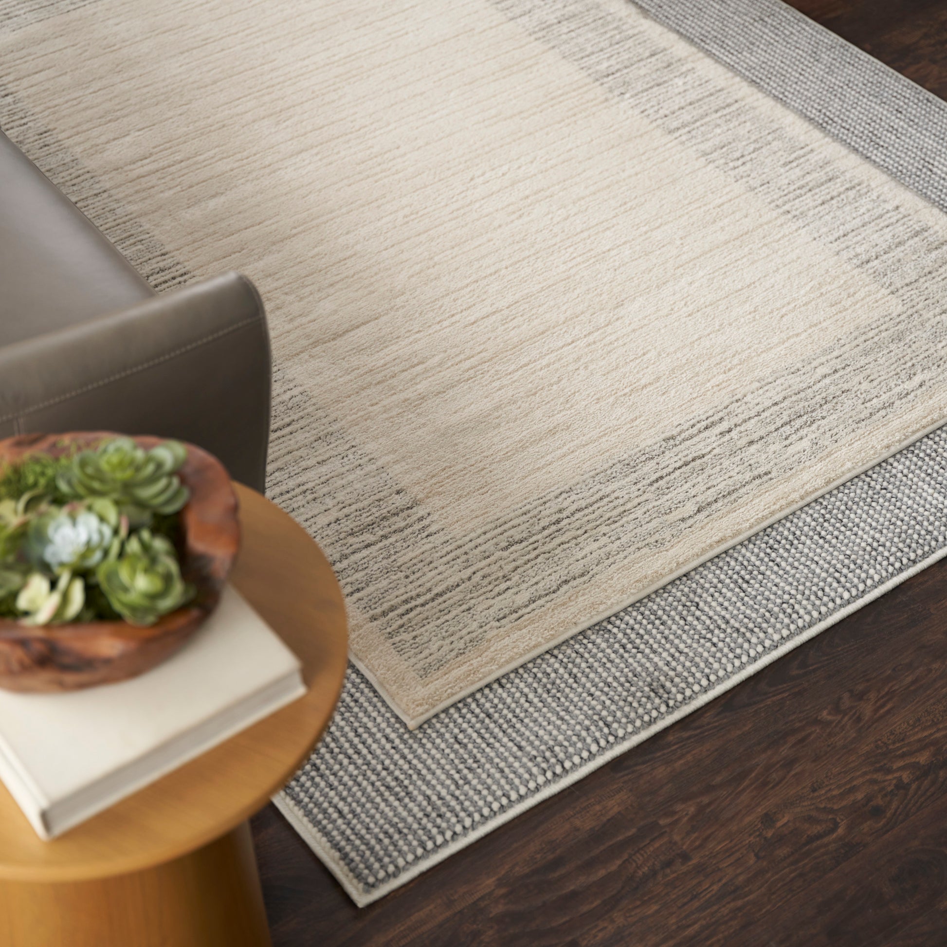 Beige textured area rug layered over a gray runner on dark wood floor, beside a round wooden side table with plant and books.