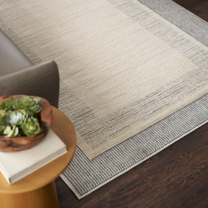 Beige textured area rug layered over a gray runner on dark wood floor, beside a round wooden side table with plant and books.