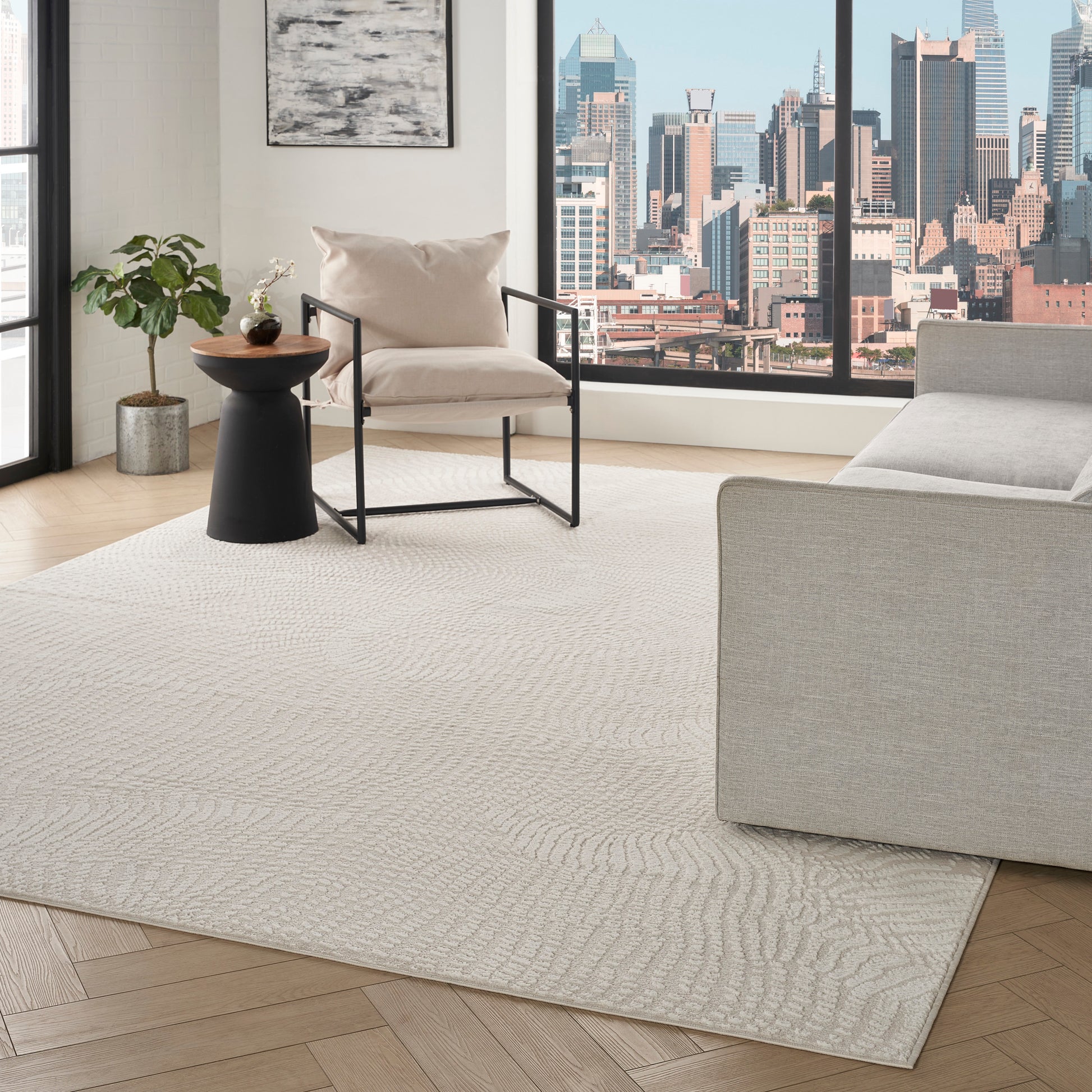 Modern living room with light beige patterned rug, beige armchair with black metal frame, gray sofa, city skyline view.