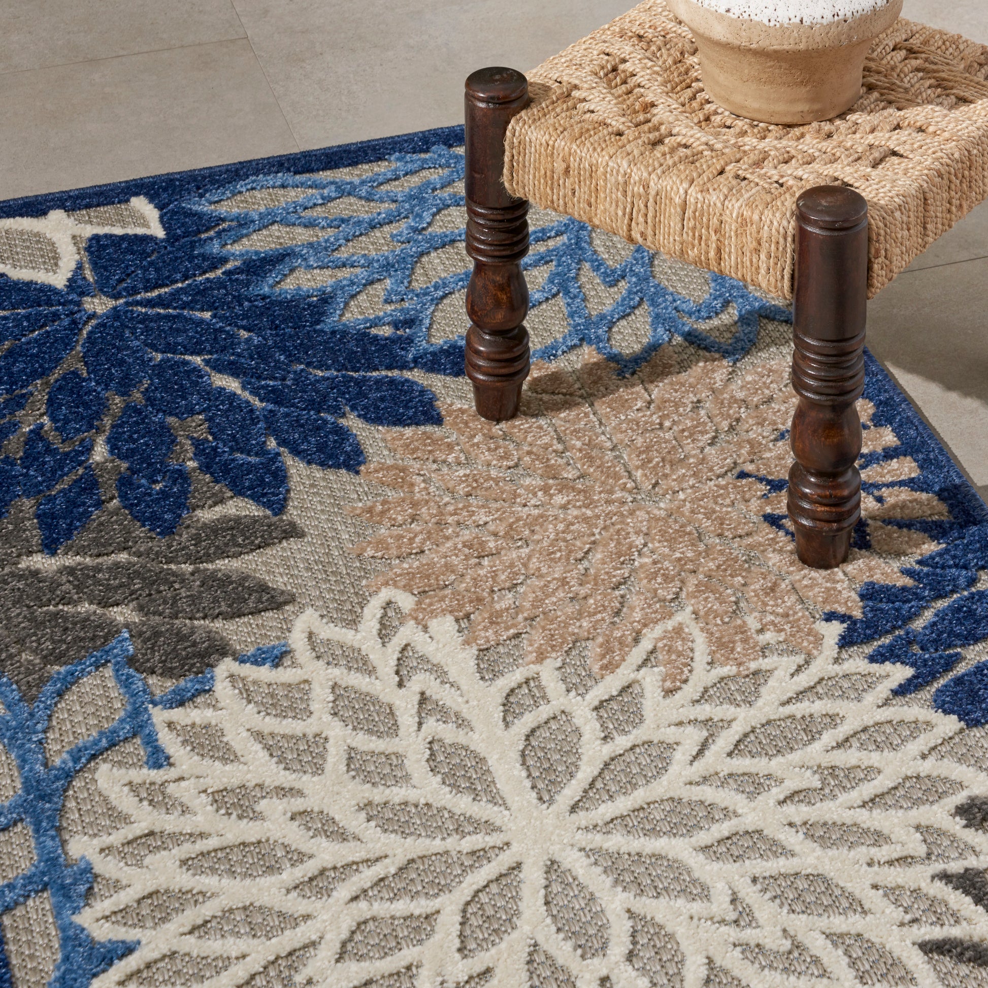 Woven area rug with a large cream floral medallion in blue and beige; dark wood‑legged stool sits on it.