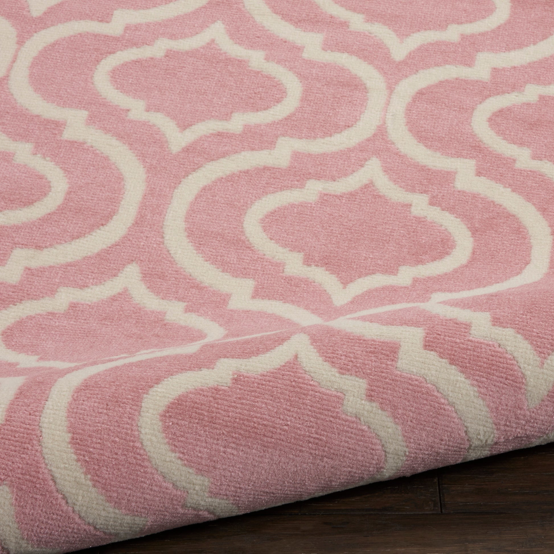 Pink rug with white trellis lattice pattern, woven texture, folded corner on dark wood floor.