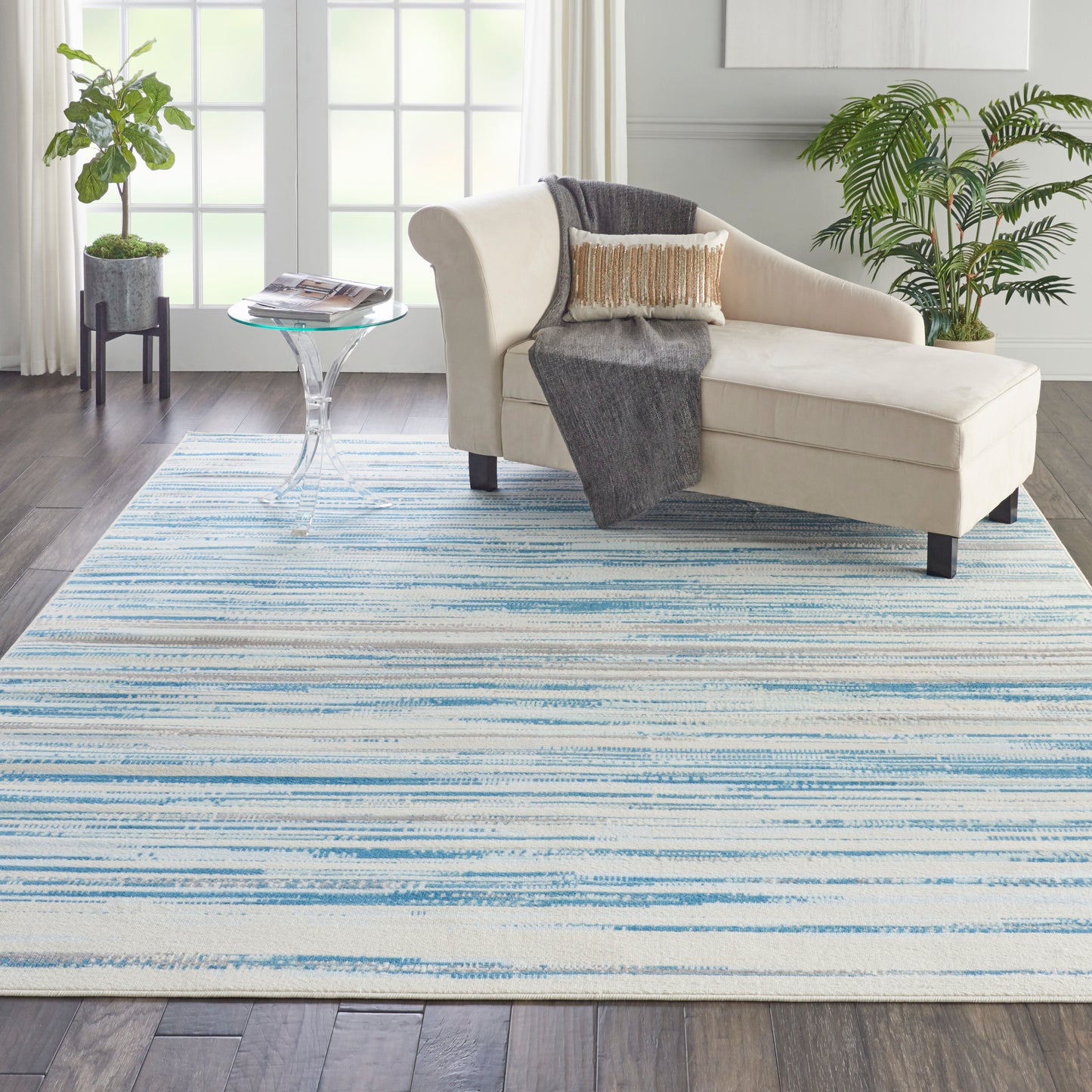 Beige chaise lounge on a blue striped area rug in a bright living room with plants and a glass side table.