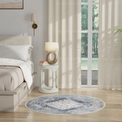 Neutral bedroom with white bedding, round white nightstand, wood-ring table lamp, blue circular rug, and sheer curtains over glass doors.