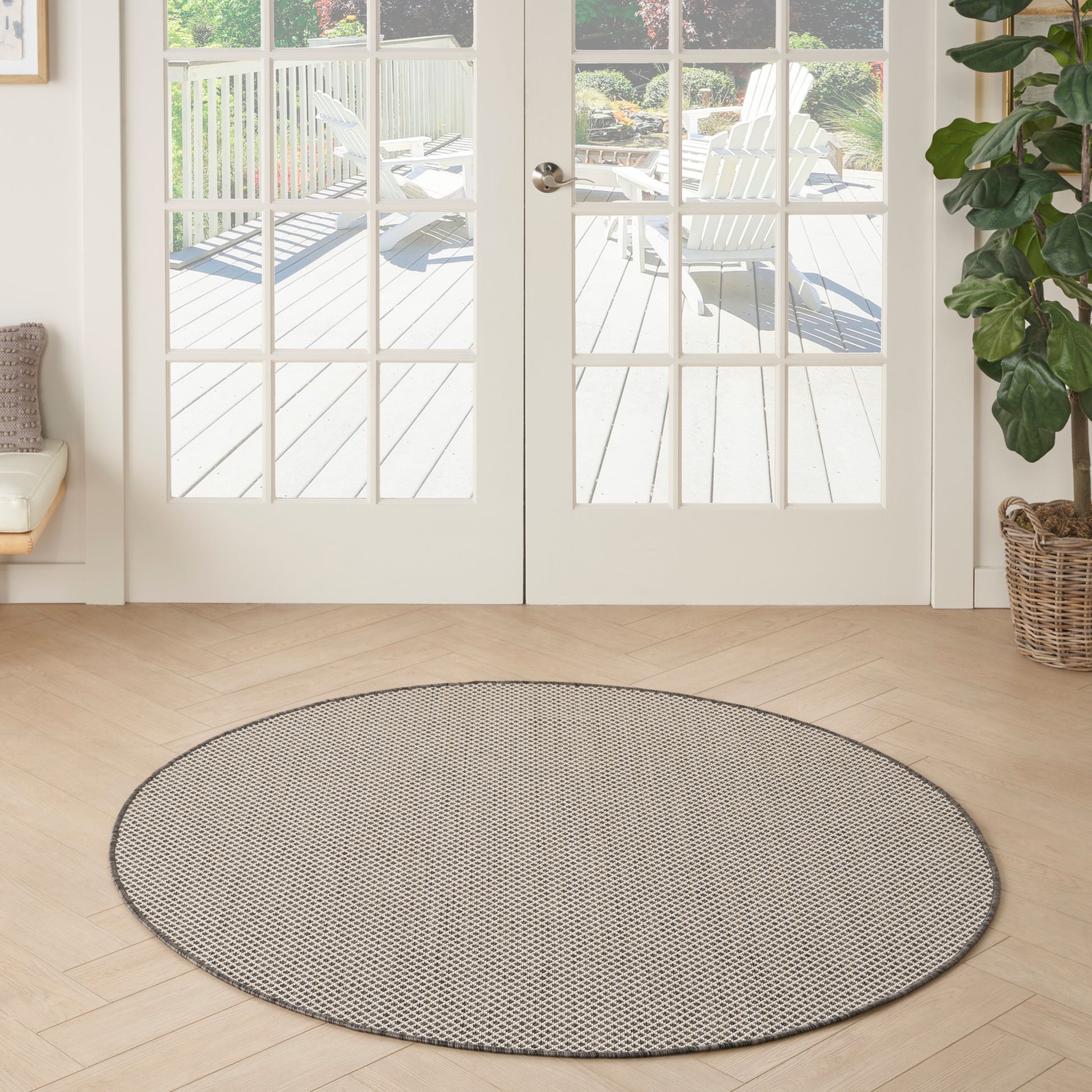 Round flat-weave rug in gray speckle on light wood floor in a bright living room with glass French doors and a potted plant.