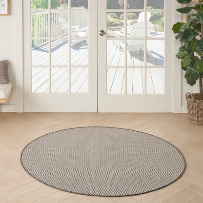 Round flat-weave rug in gray speckle on light wood floor in a bright living room with glass French doors and a potted plant.